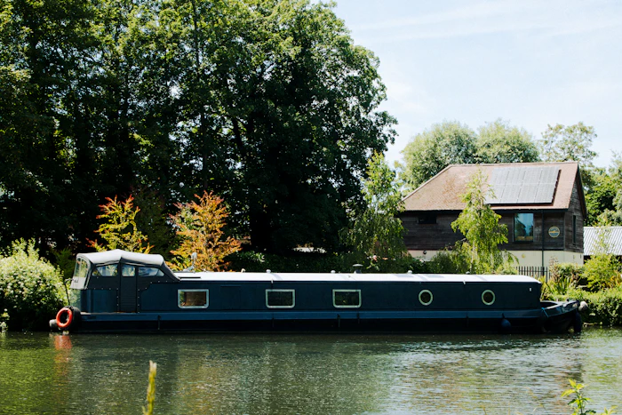 A dark blue canal boat on still water with trees and a house on the bank