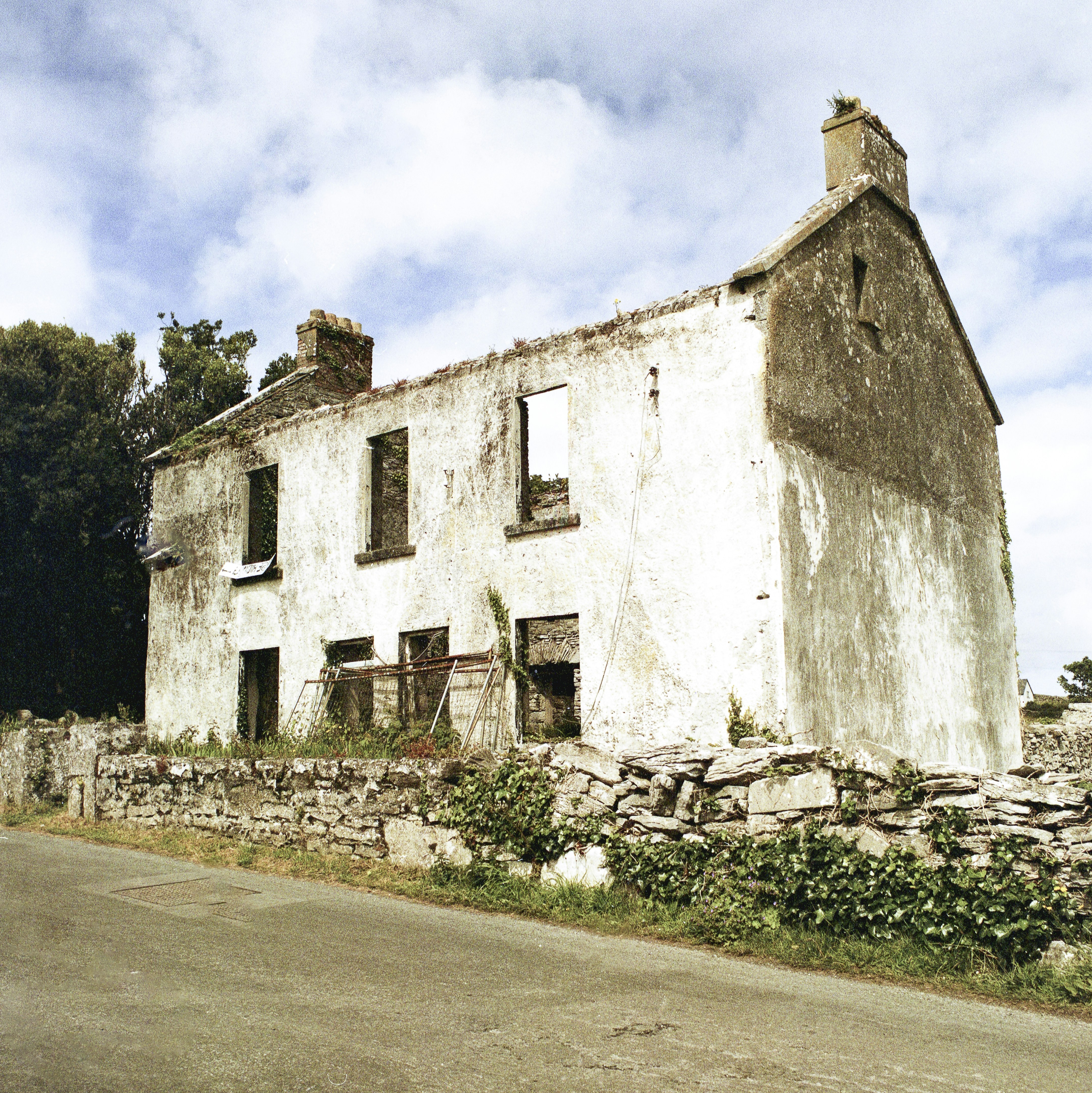 Dilapidated white house with empty windows and a stone wall.