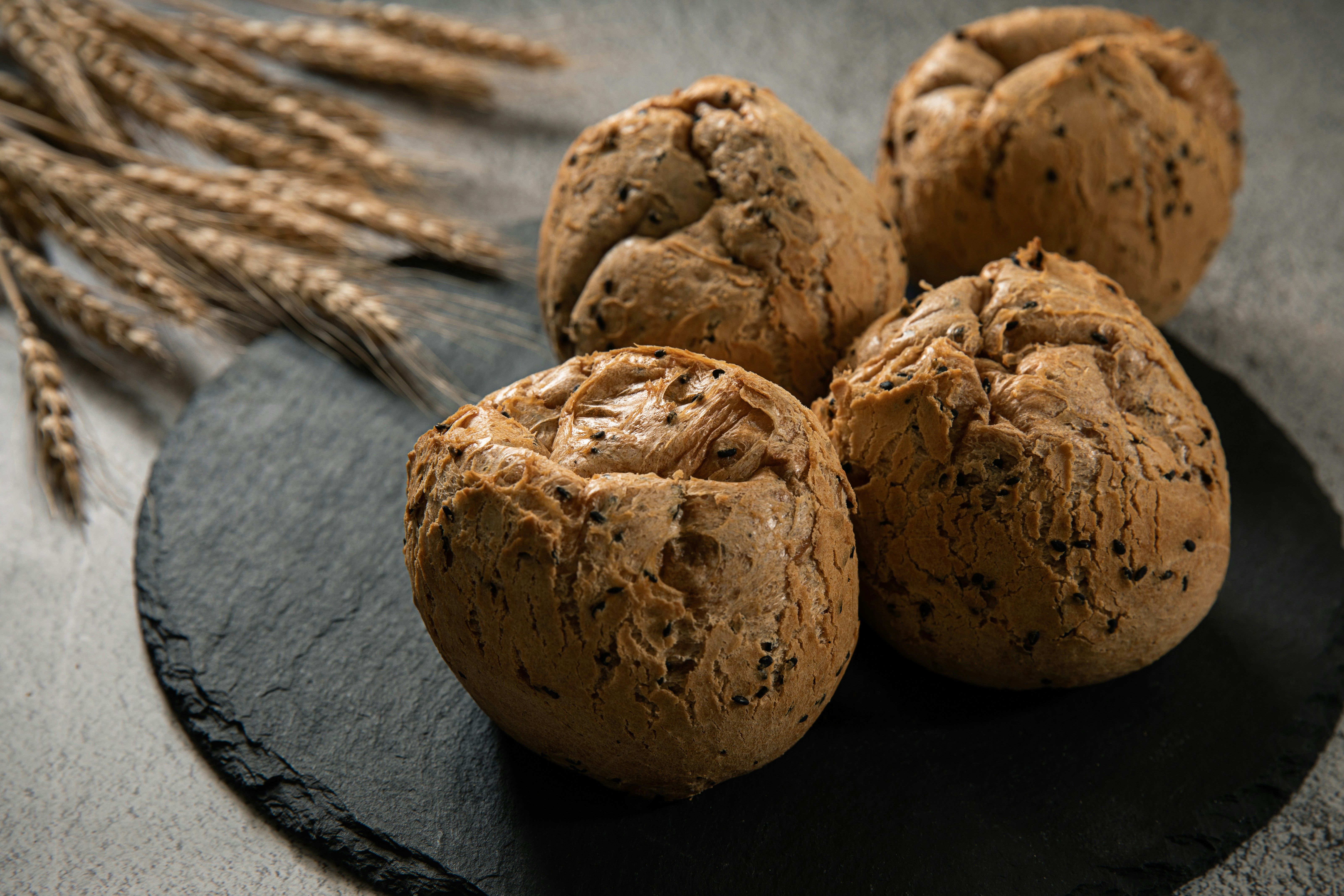Four dark bread rolls on a slate board with wheat.