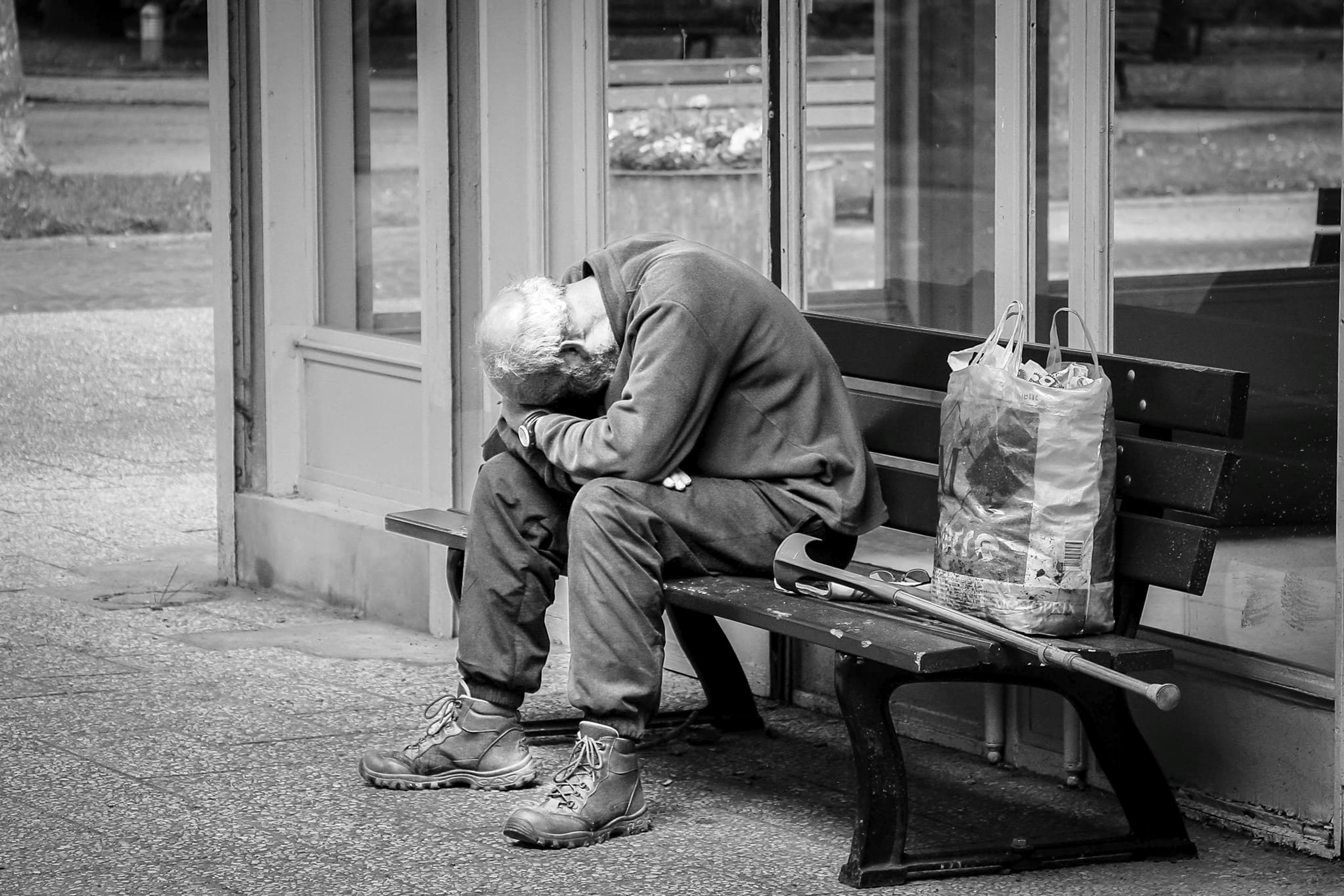Elderly person slumped on a bench with a walking stick.