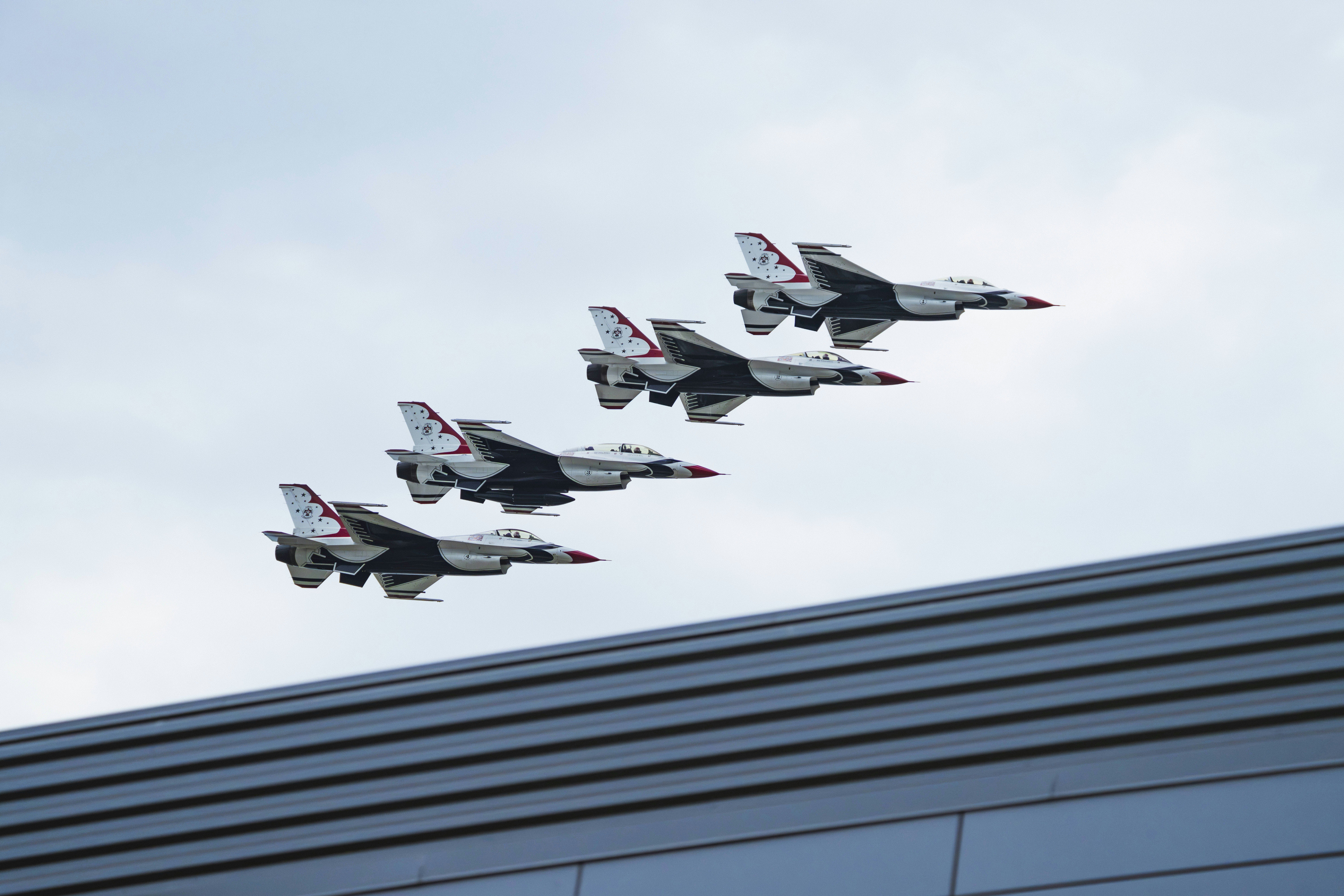 5機の戦闘機が編隊を組んで近代的な建物の上空を飛行しています