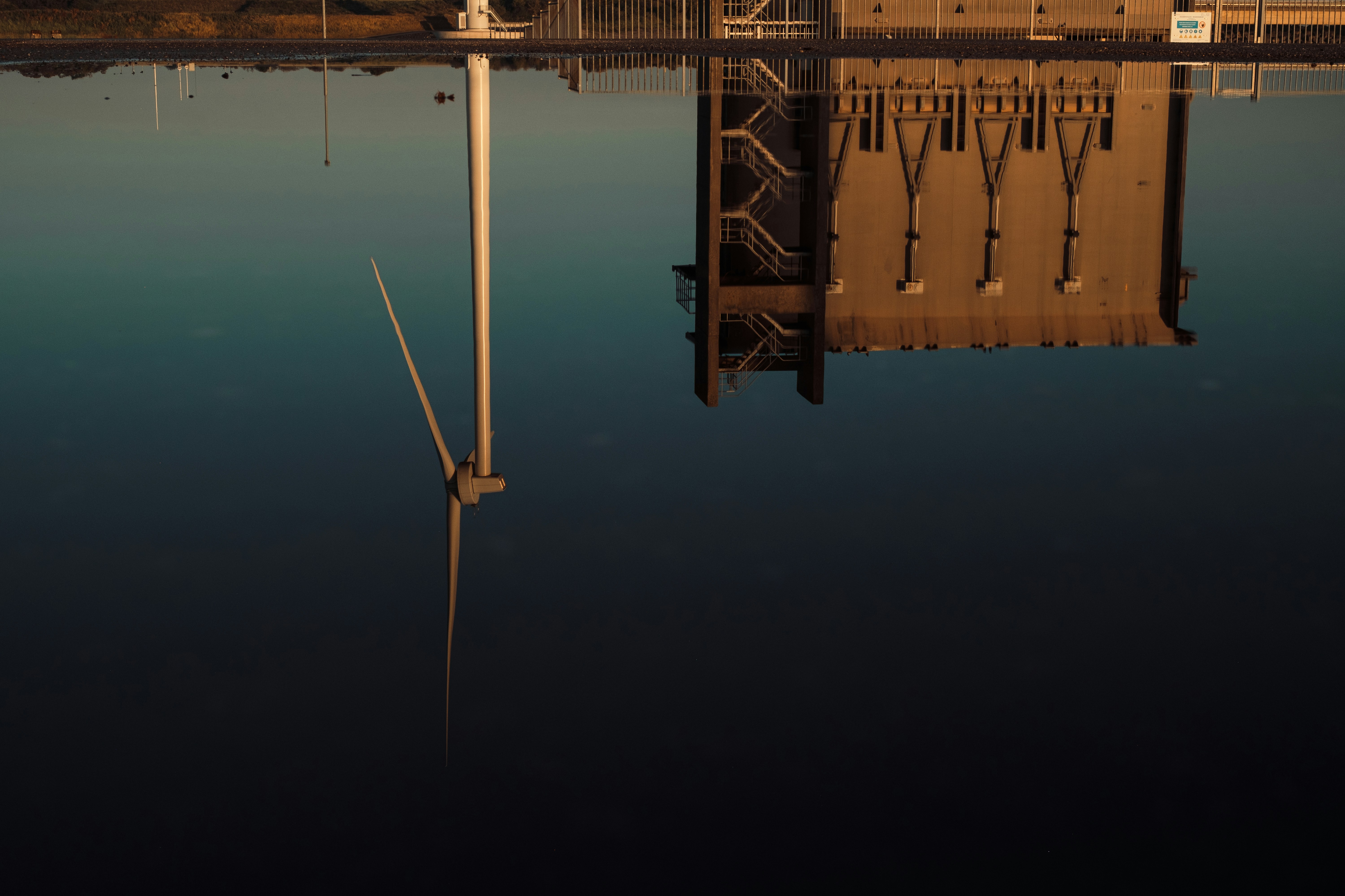 Technology A Near Perfect, Glass Like Reflection Of A Wind Turbine And A Massive Industrial Storm Surge Barrier Structure In Perfectly Still Water. The Warm Golden Hour Light Illuminates The Concrete And Steel, Contrasting Beautifully Against The Deep, Gr
