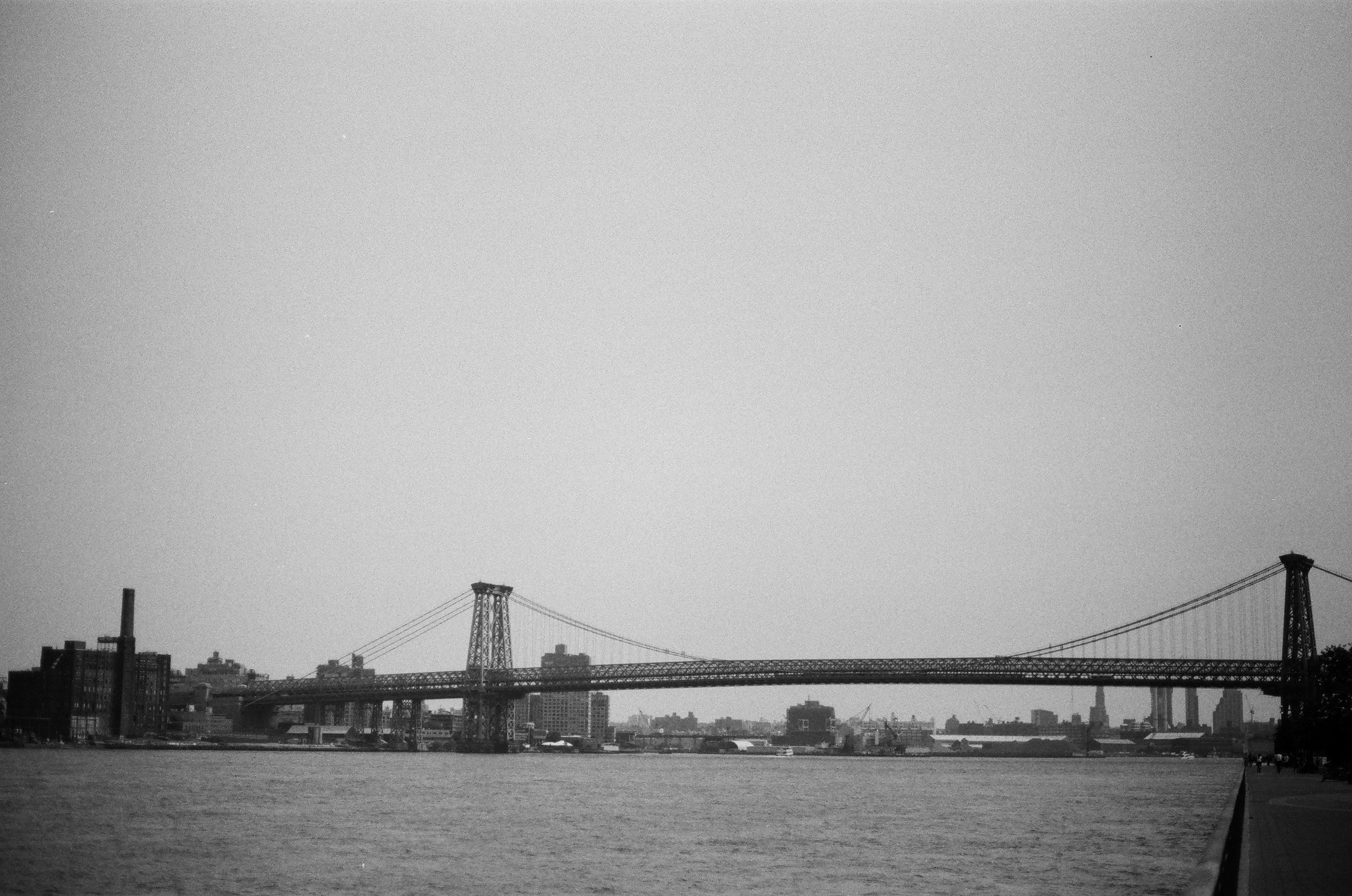 A grayscale suspension bridge over water with city buildings.