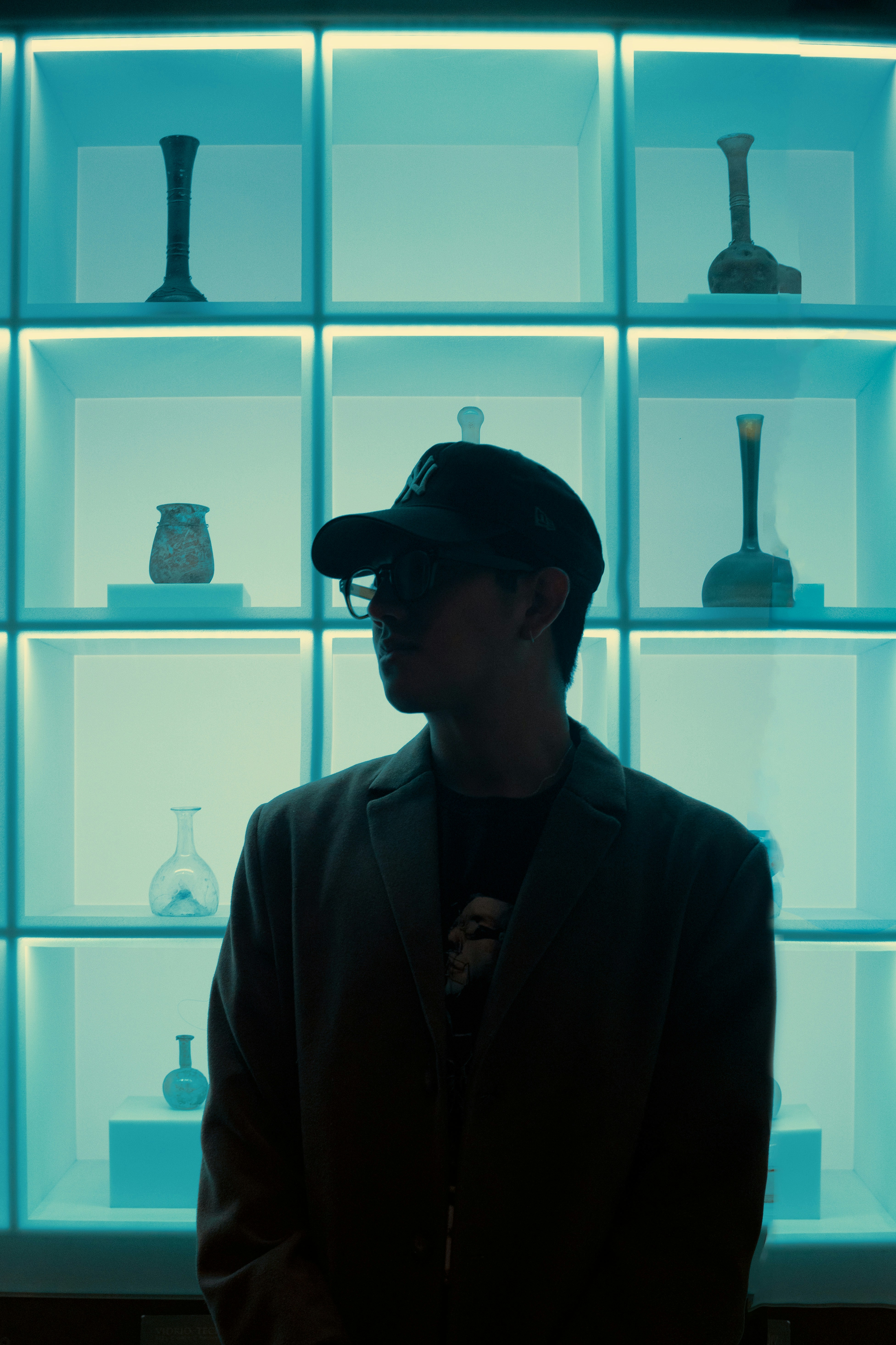 A person in silhouette stands before illuminated shelves displaying vases.
