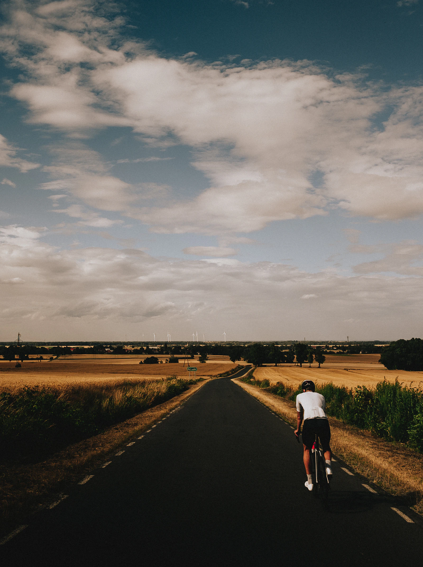 A lone cyclist rides away down a straight road cutting between harvested fields under broken cloud