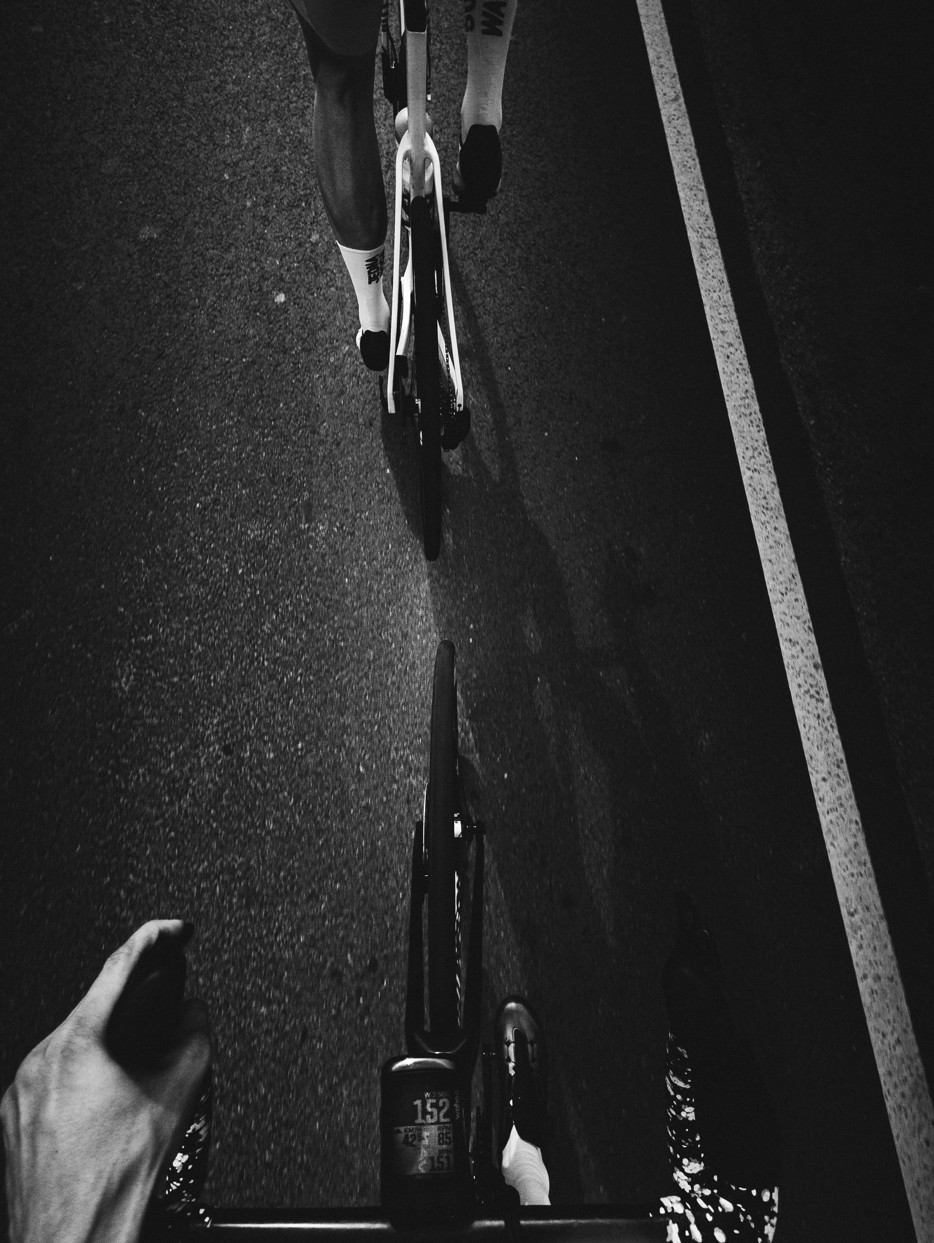 A cyclist rides behind another on a dark road.