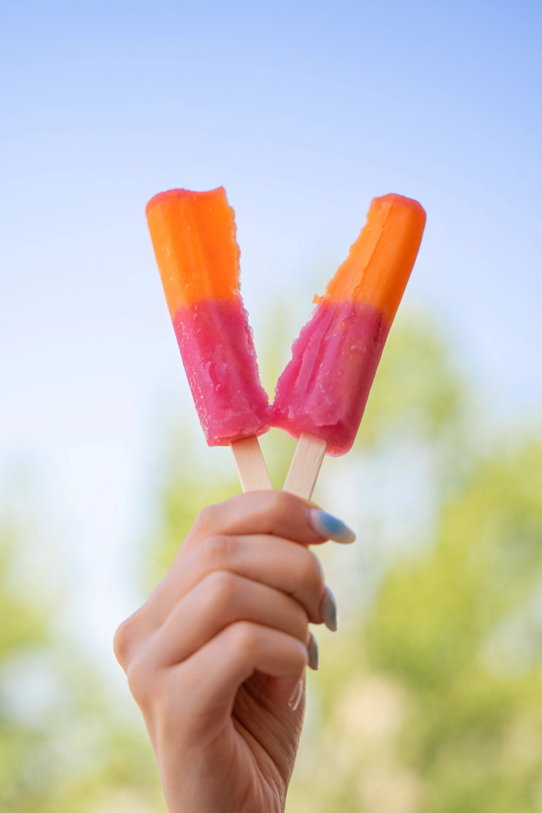 A hand holding up two orange and pink layered pops against a blue sky