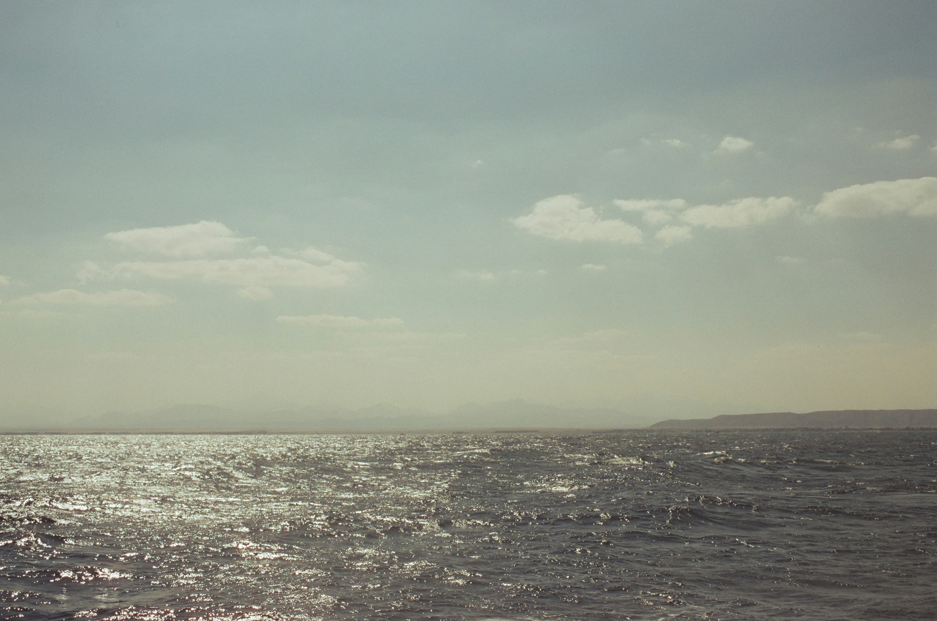 Ocean Beautiful 35Mm Film Seascape Showing A Wind Rippled Dark Ocean Surface Sparkling With Sunlight Glinting Off The Waves In The Foreground, With A Hazy Mountainous Coastline Running Along The Right Horizon And Dramatic Billowing White Clouds Above A Pa