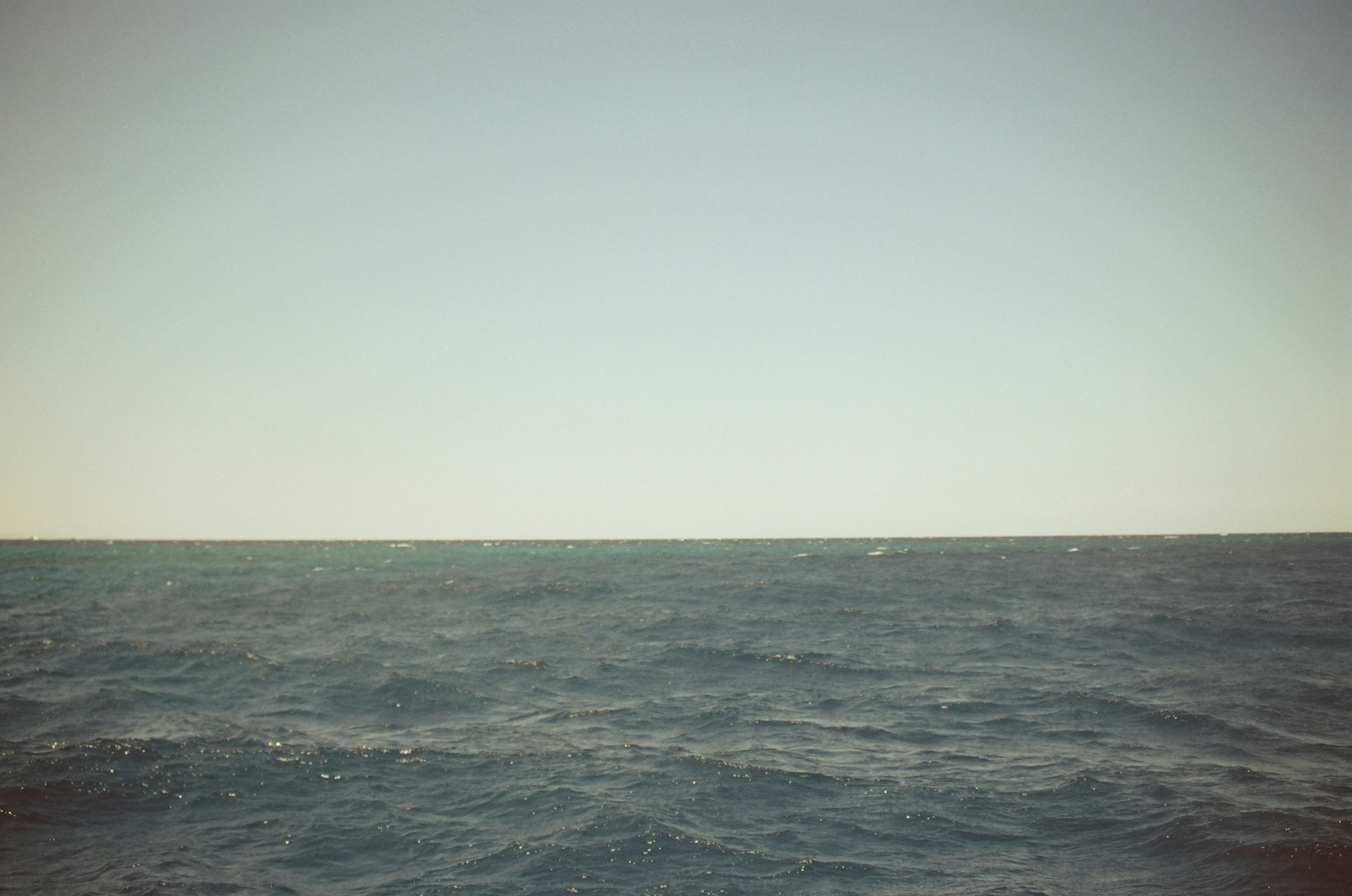 Space Stark Minimalist Seascape Captured On 35Mm Film, With Dark Blue Gray Choppy Water Filling The Lower Half Of The Frame And A Warm Pale Sky Above, Meeting At A Narrow Horizon Band Where A Sliver Of Turquoise Water Catches The Light. Almost Abstract In