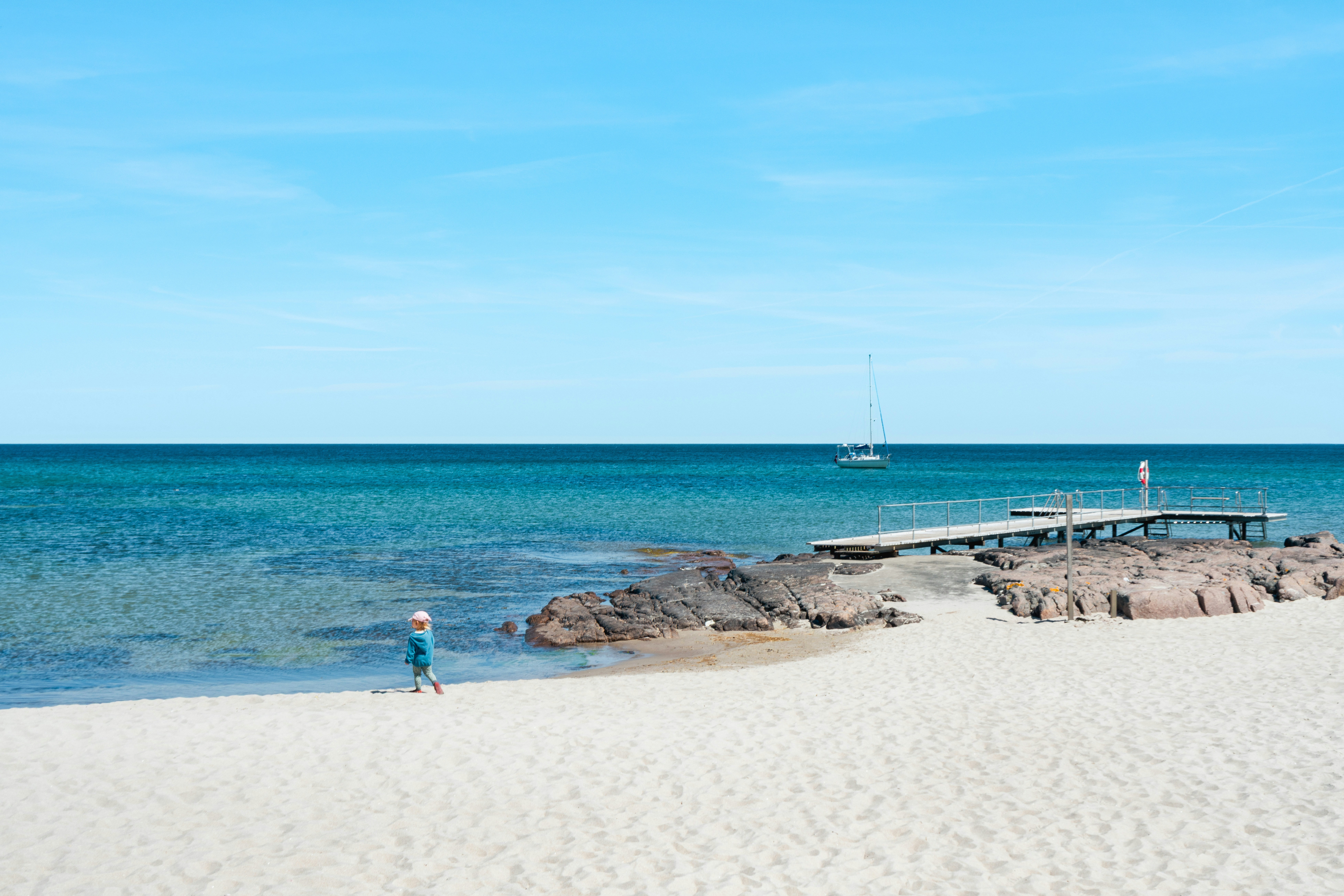 Niño en una playa de arena con muelle, rocas y velero.