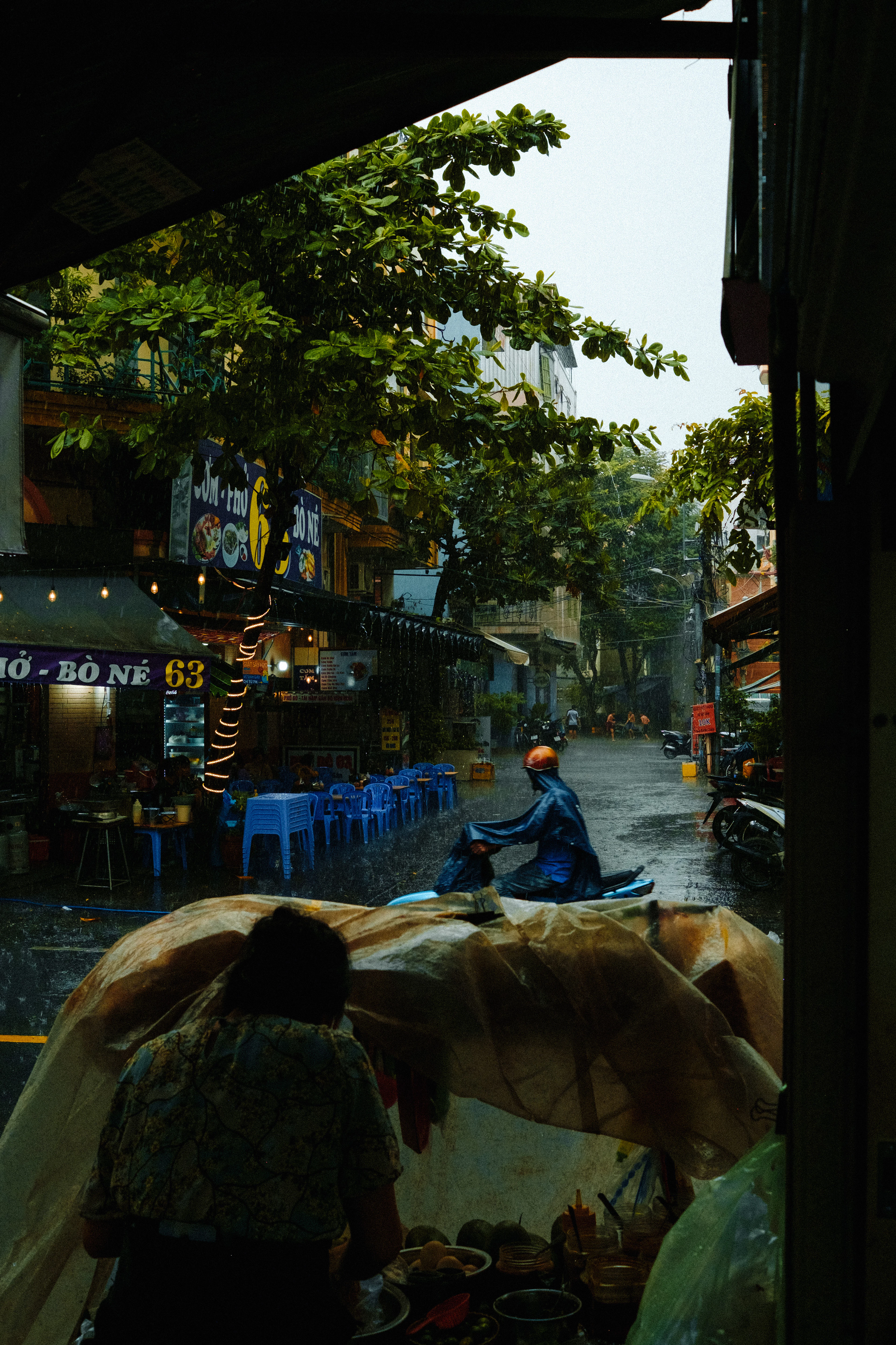 A tropical downpour seen from inside a Ho Chi Minh City food stall, a rider in a poncho sliding past rows of empty blue plastic stools and the lit sign of a pho and bo ne shop across the wet street.