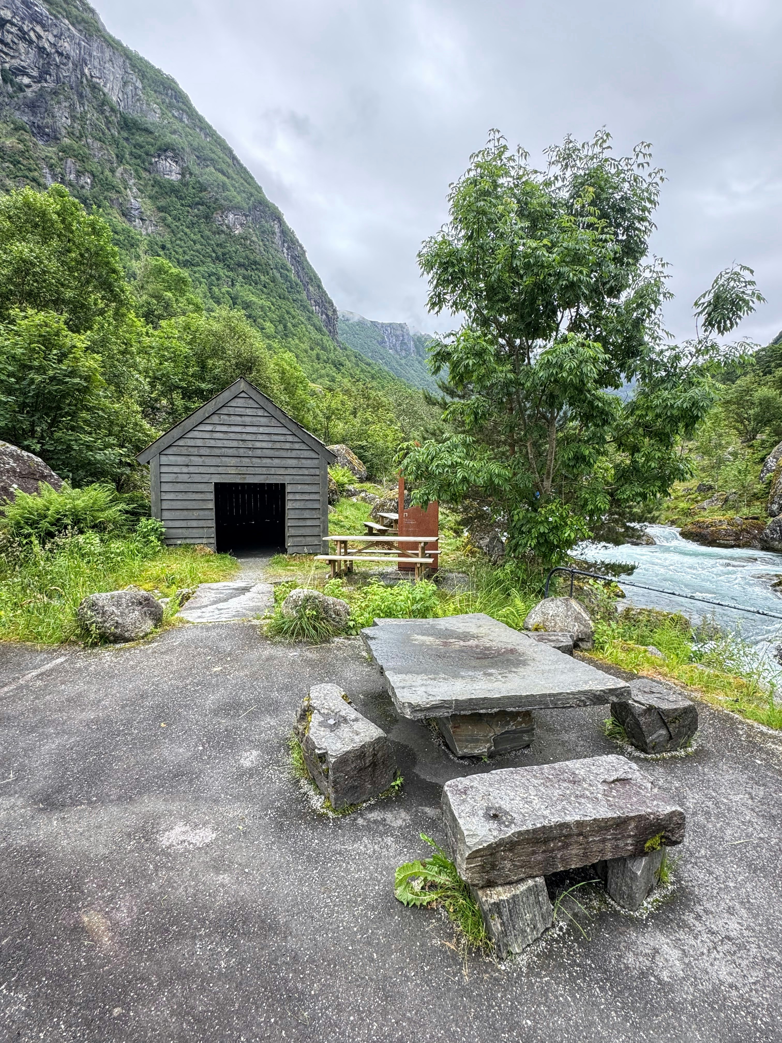 Cabaña rústica y mesa de picnic de piedra junto a un río de montaña.