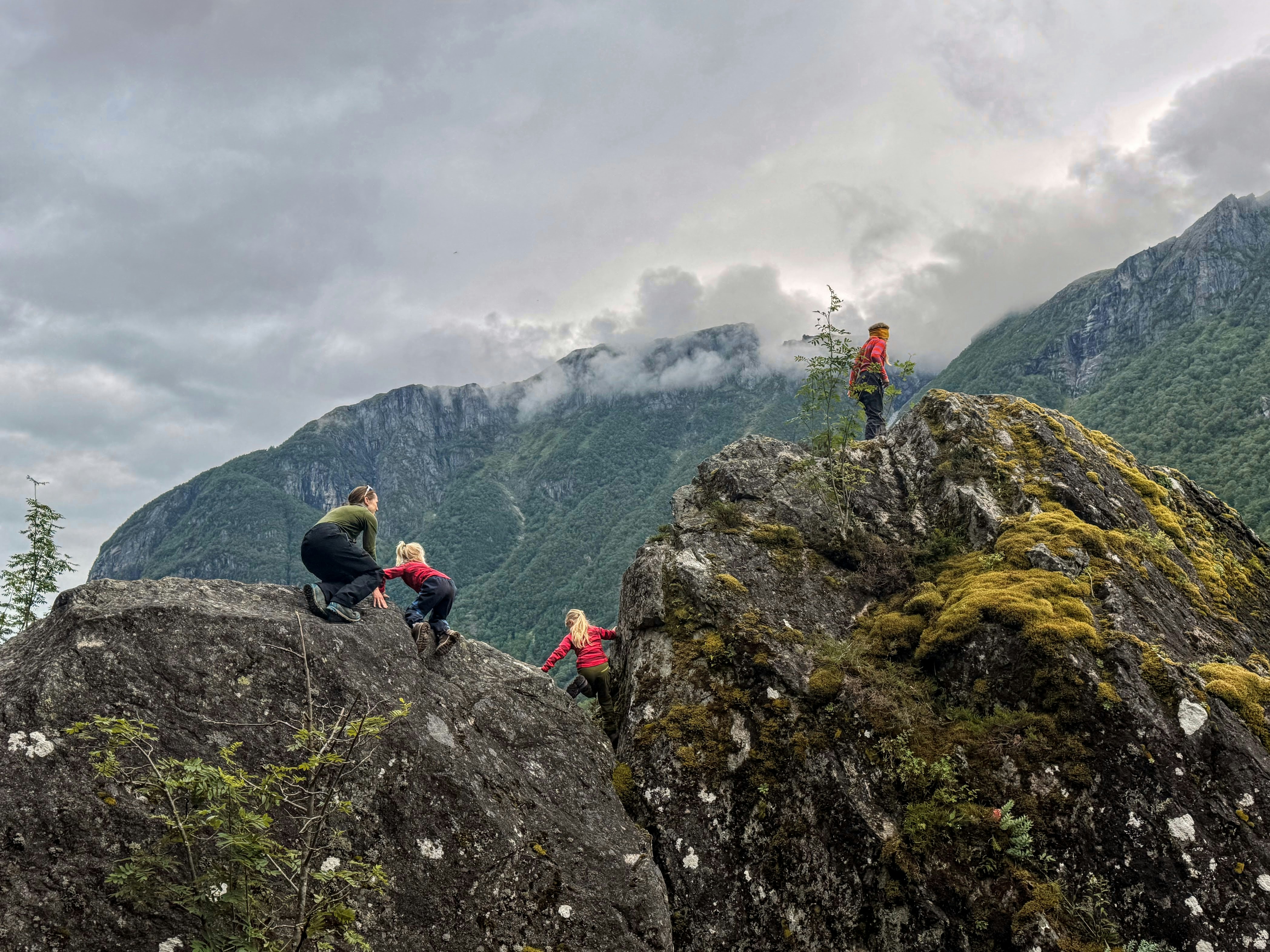Gente escalando rocas cubiertas de musgo con montañas y cielo nublado.