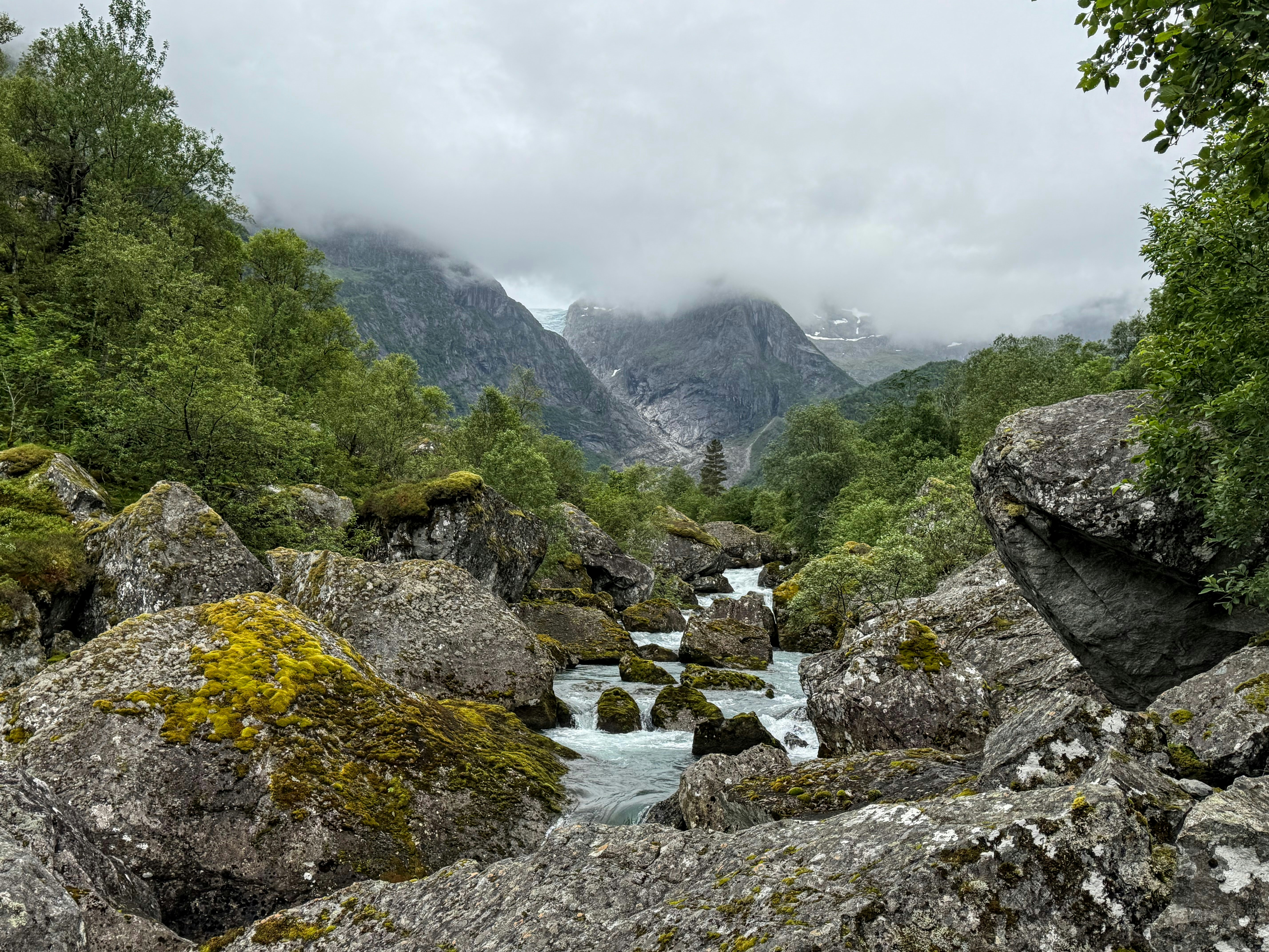 Un río sereno fluye entre rocas cubiertas de musgo con un fondo nublado de montaña.