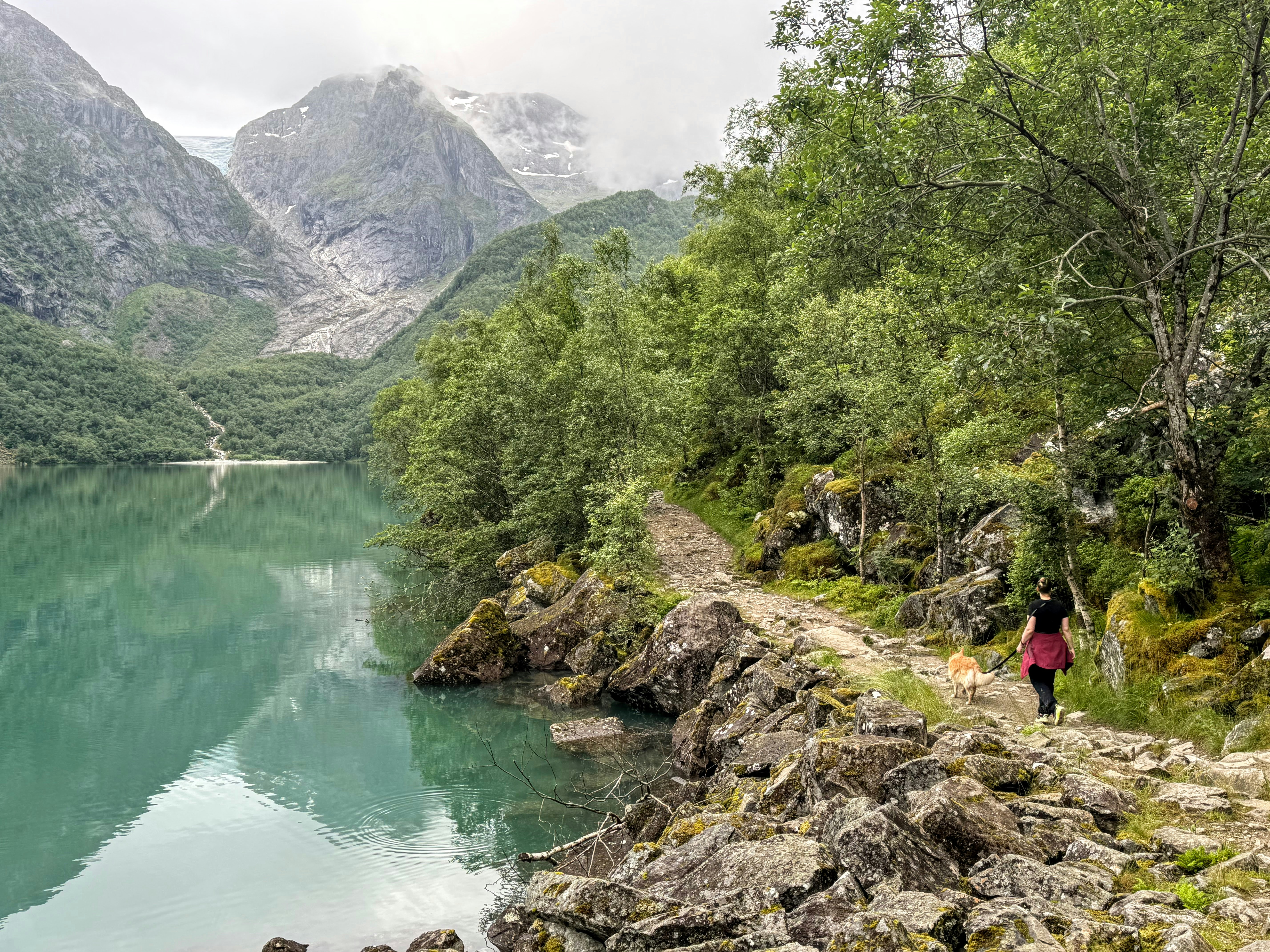 Mujer camina por un sendero rocoso junto a un lago de montaña tranquilo.