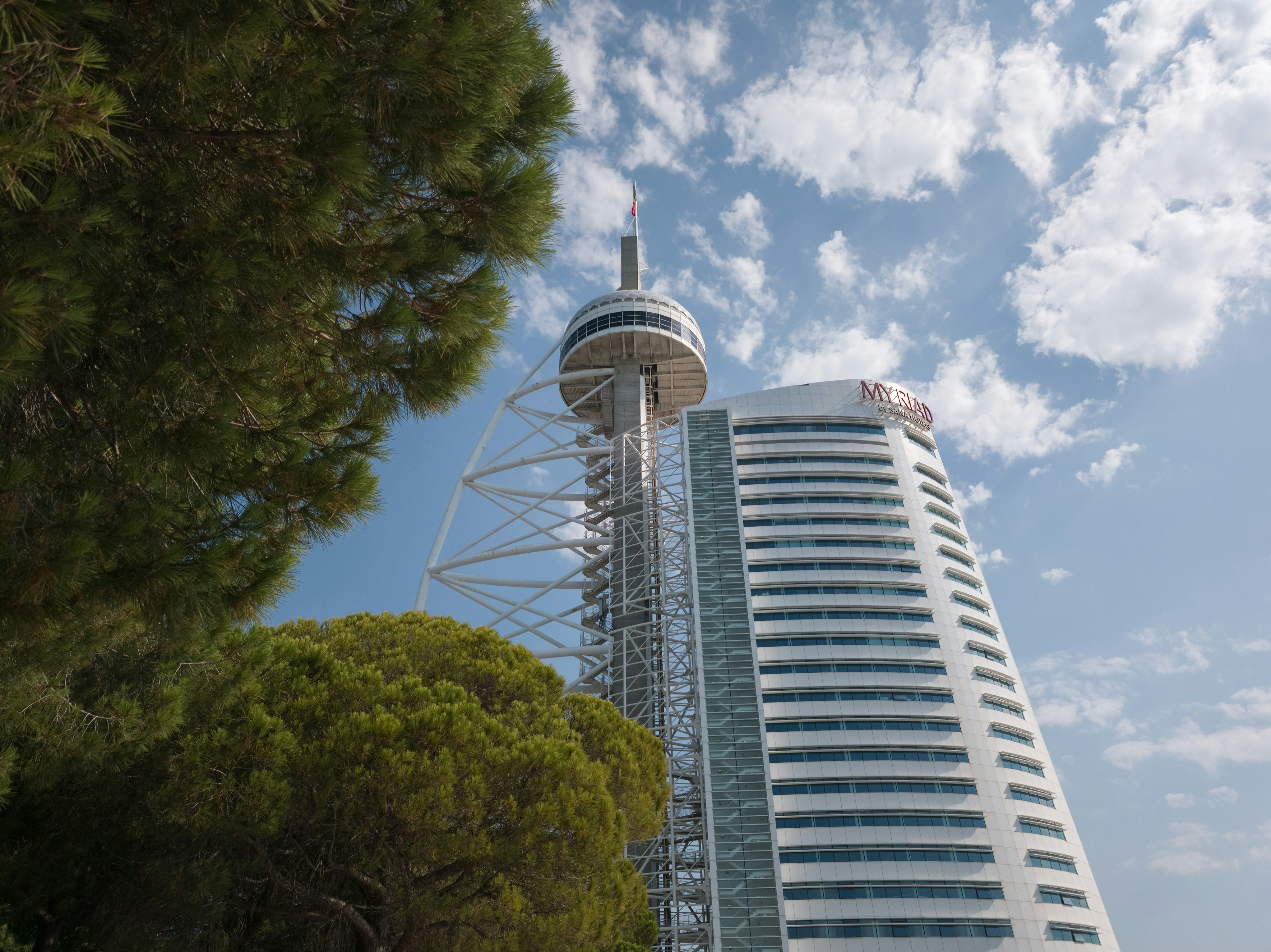 Modern tower and building framed by green trees.
