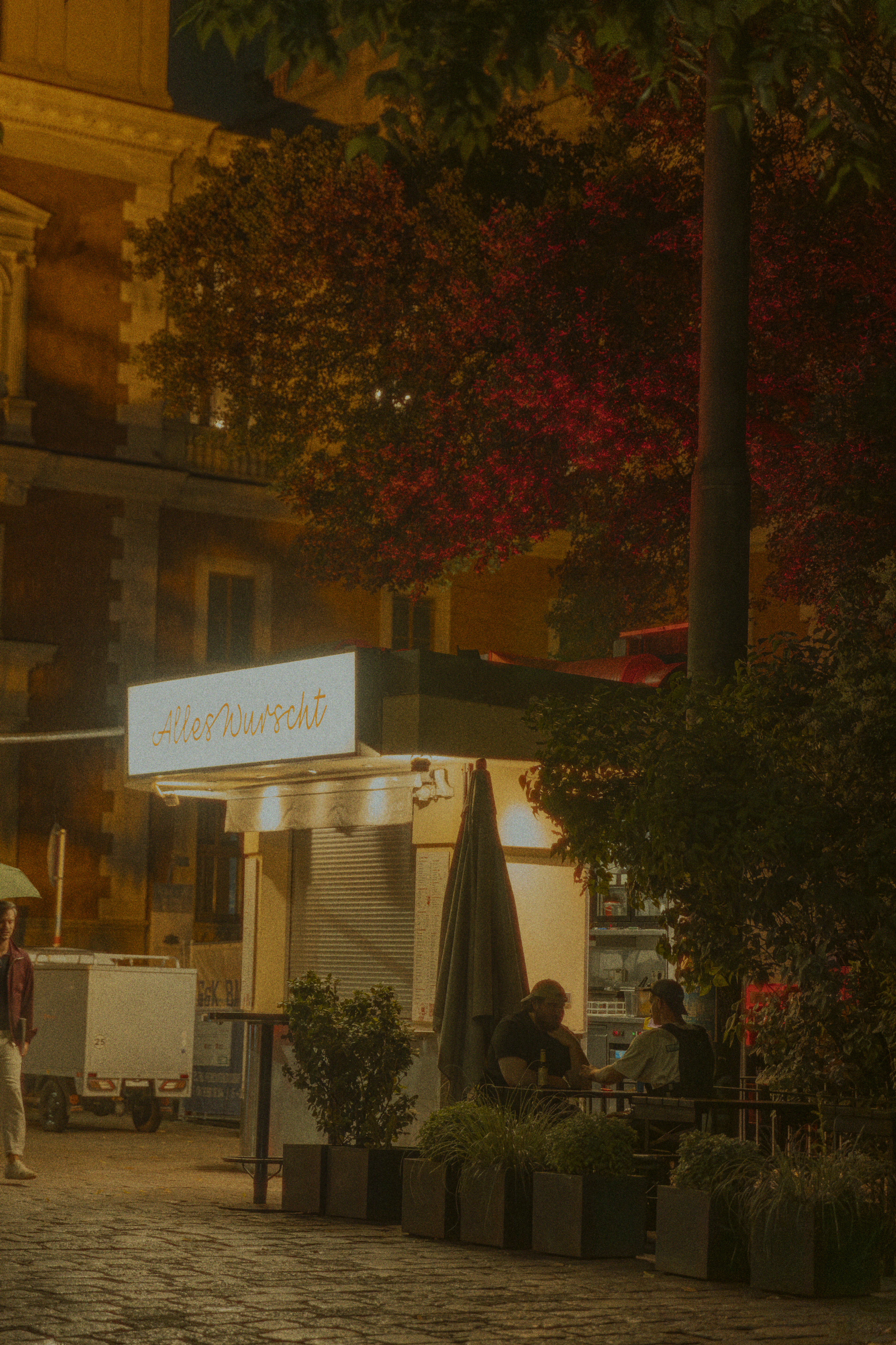 A lit sausage kiosk in Vienna at night, its white sign reading 'Alles Wurscht' glowing under a tree in deep red autumn leaf, two people talking at a bench beside planters on the cobbles and a lamplit stone facade behind.