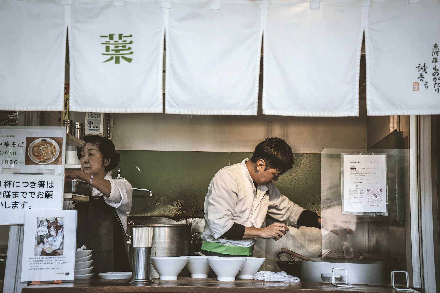 Two cooks behind a small ramen shop counter, with stock pots on the stove and a row of empty bowls ready