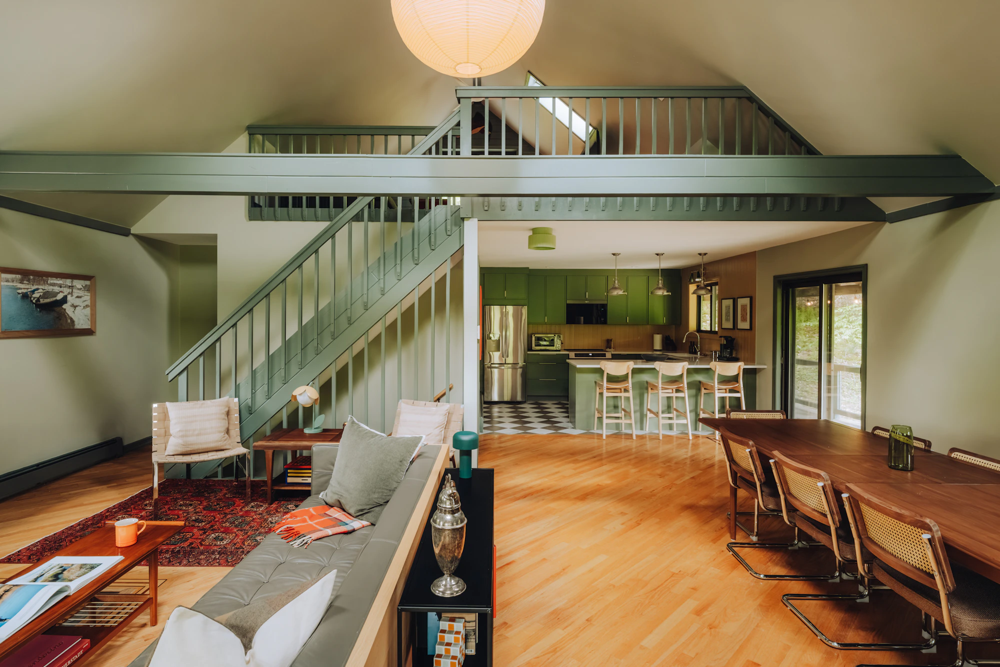 Open-plan home interior with a sage-green staircase and mezzanine railing, living area, timber dining table, green kitchen cabinetry and warm timber floors.