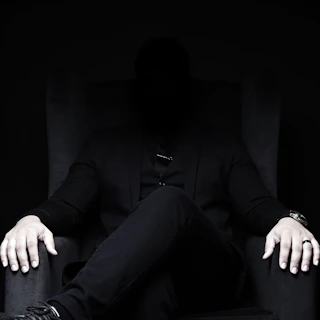 Mysterious man in dark suit sits in a chair.