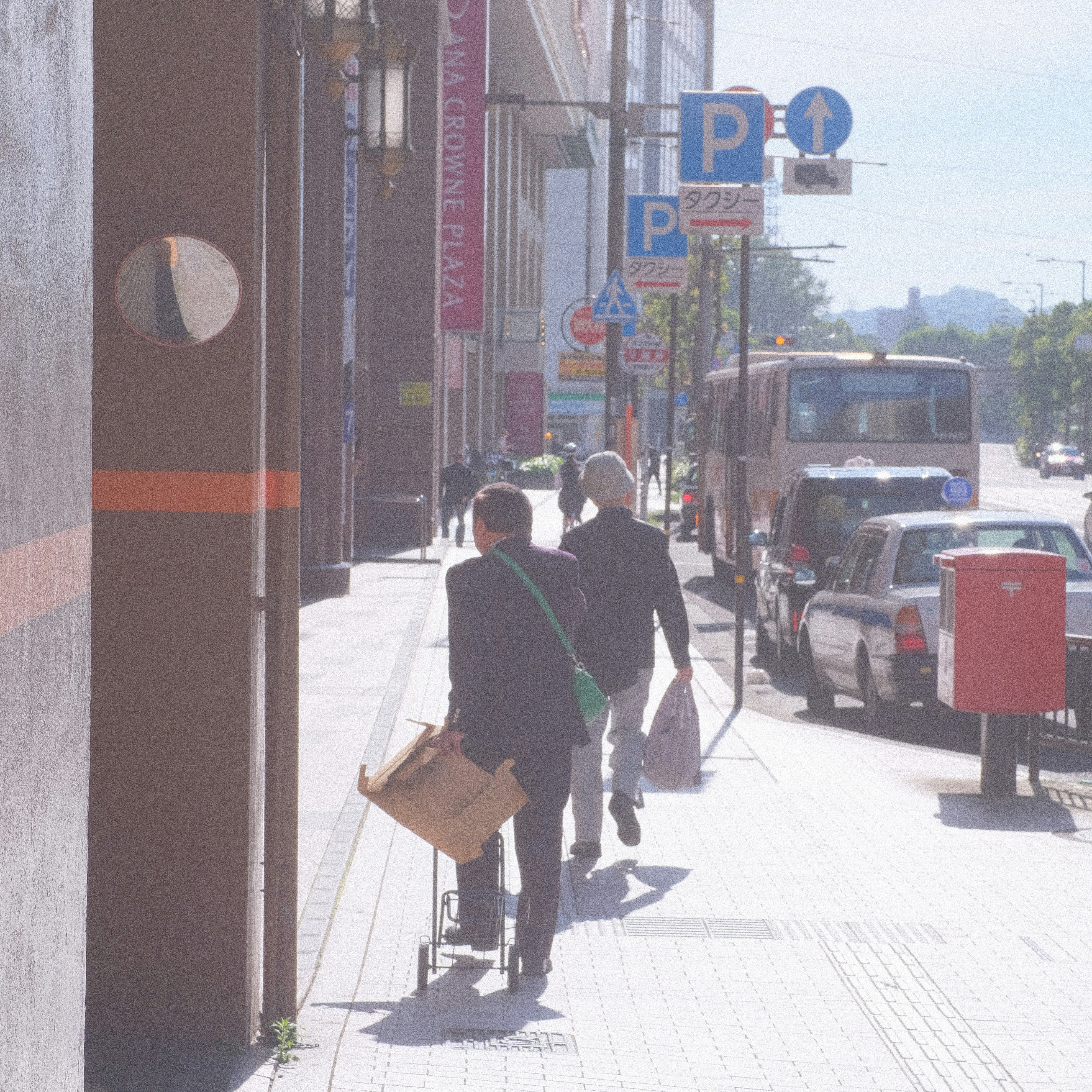 Pedestres caminham por uma calçada urbana ensolarada com veículos estacionados.