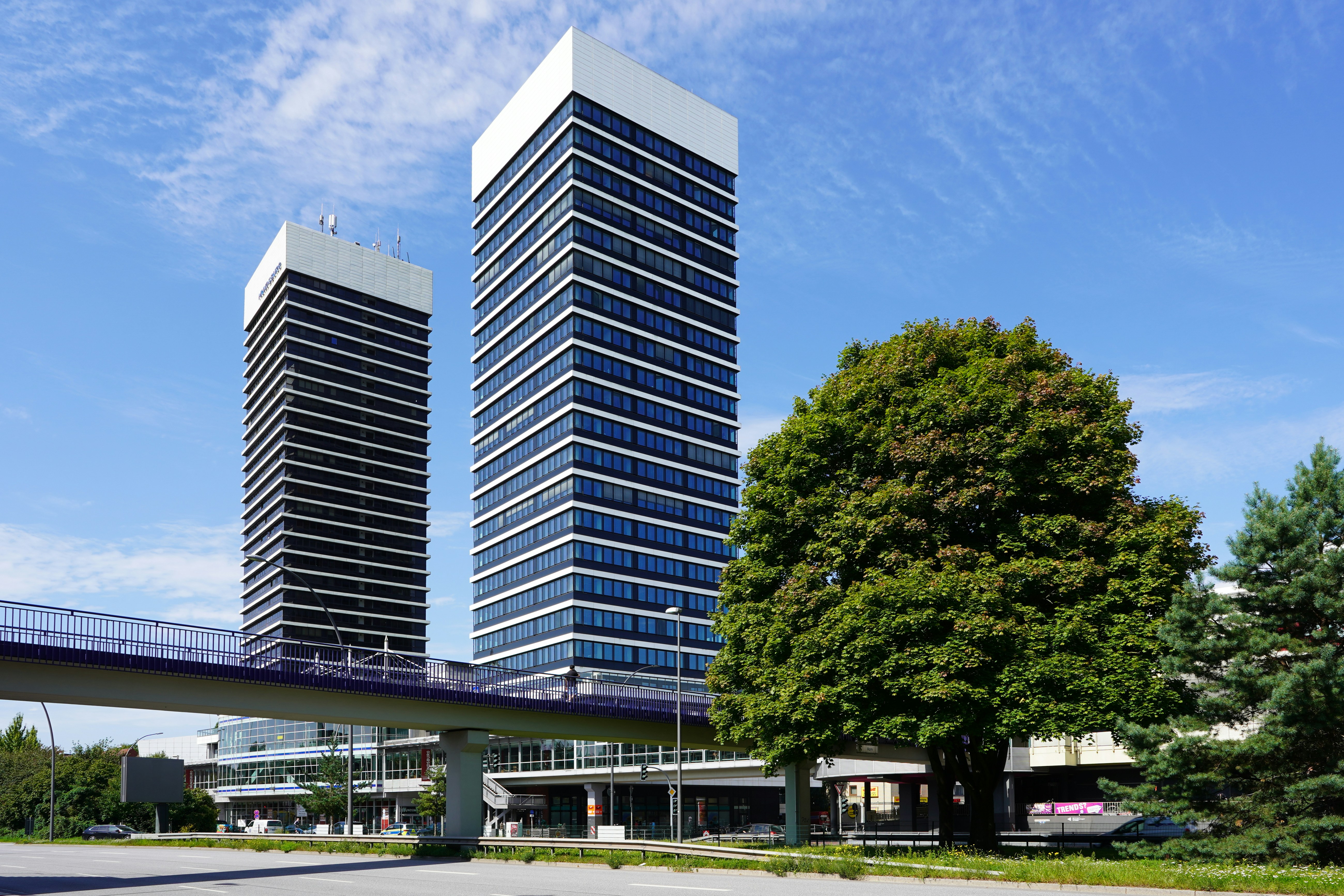 Modern buildings, pedestrian bridge, and green trees under blue sky.