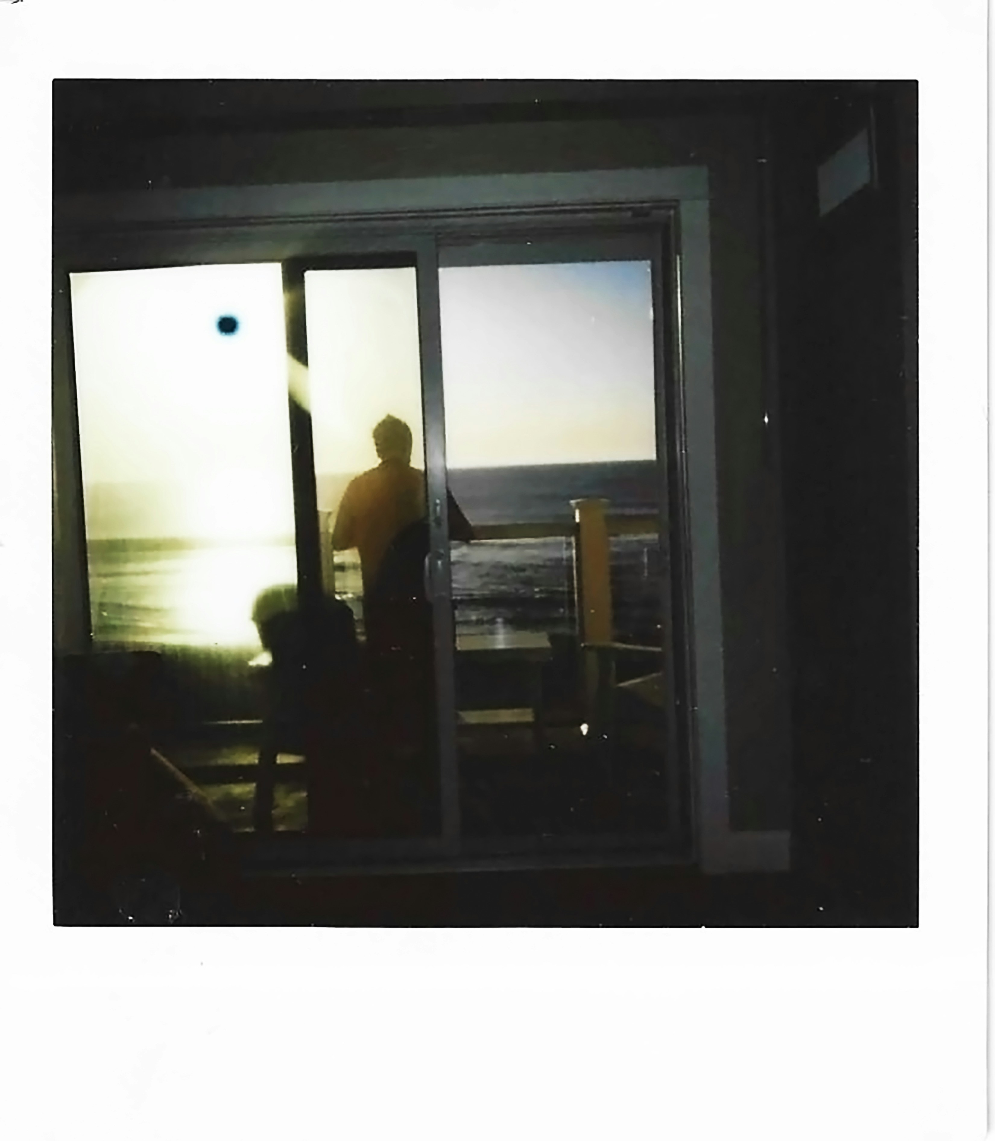 Personne en silhouette regardant l’océan se coucher de soleil à travers une porte vitrée.