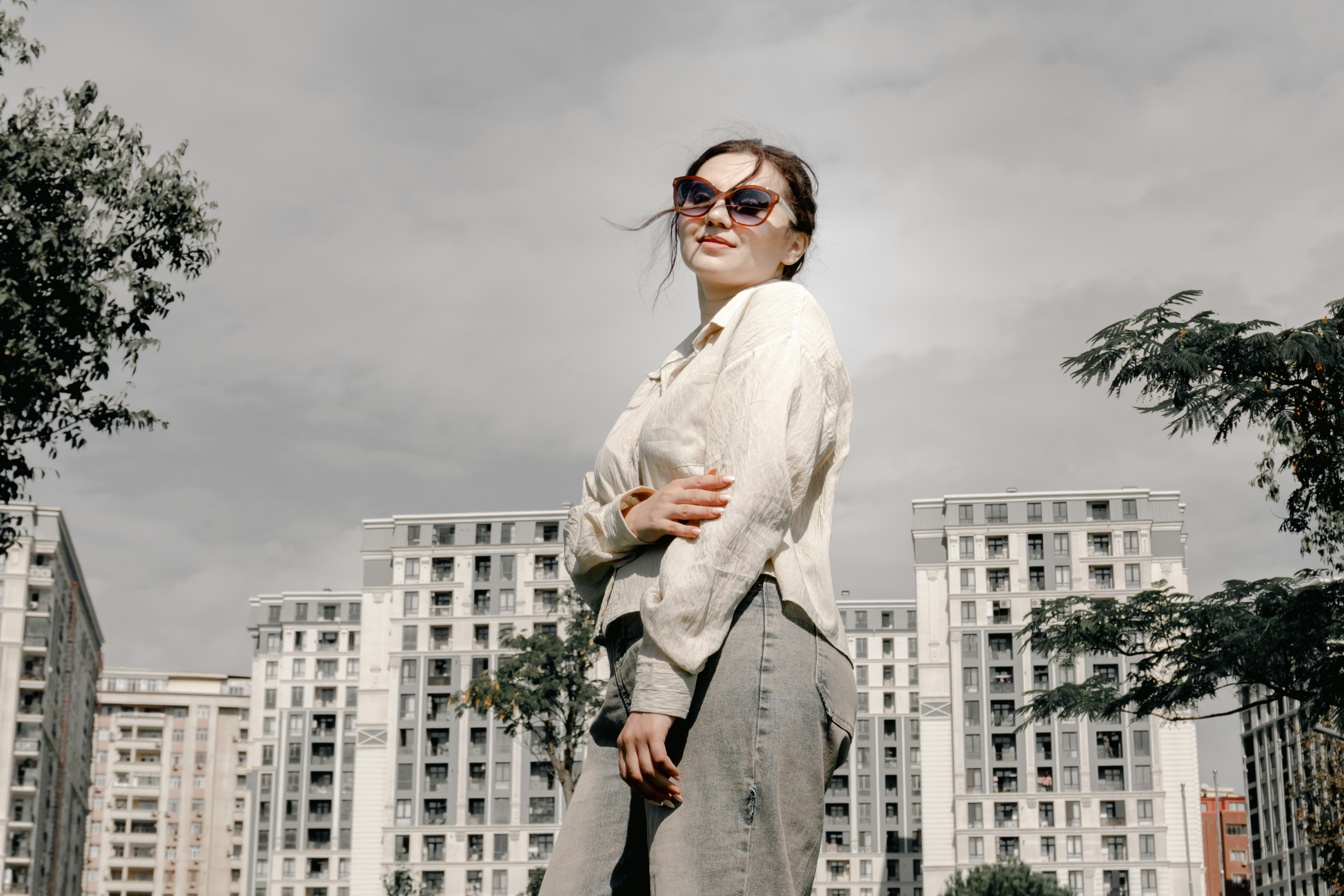 A young woman in sunglasses stands against urban buildings.