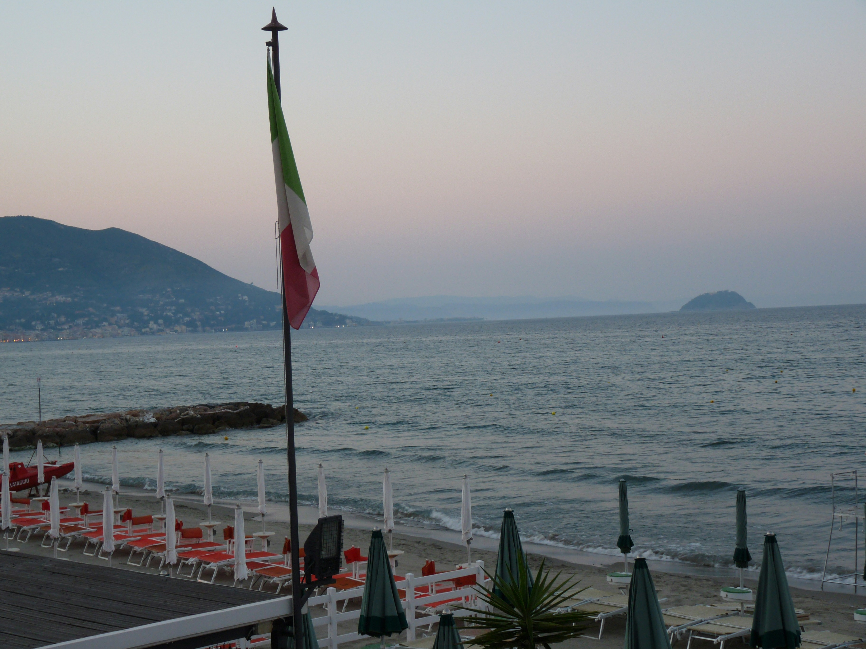 Italian flag, beach chairs, ocean, and distant mountains at dusk.