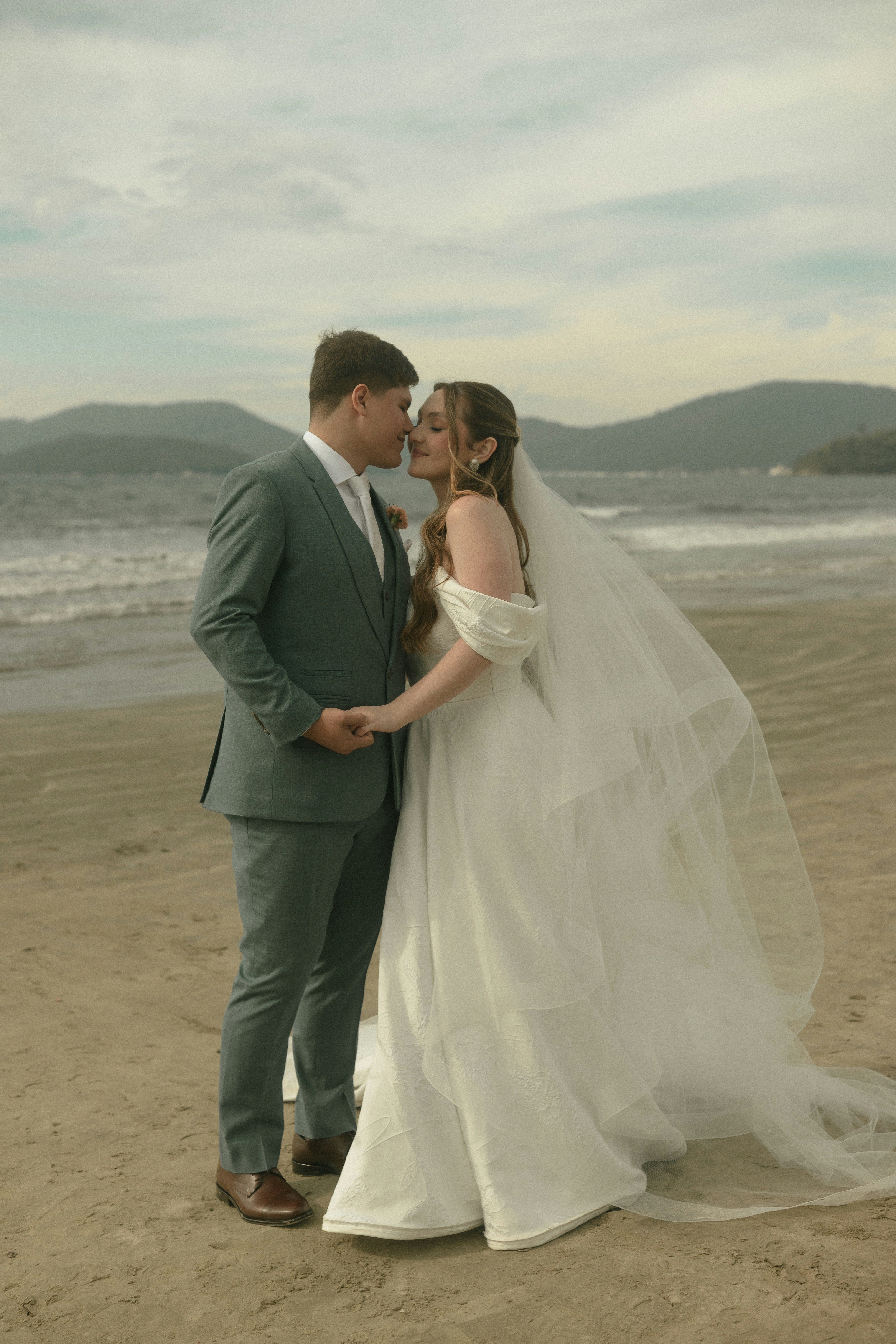 Novios besándose en una playa de arena con montañas.
