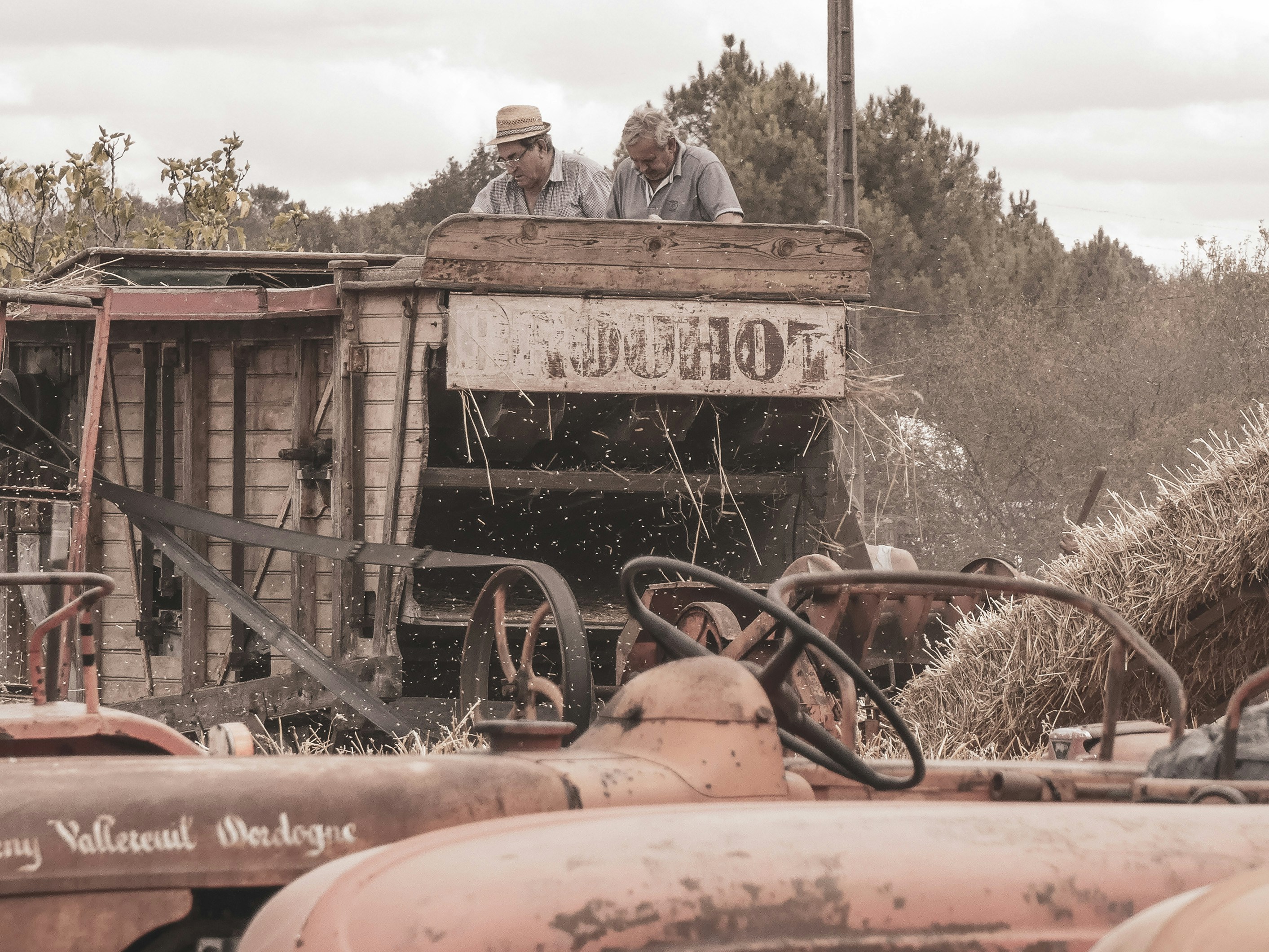 Two men operate vintage farm machinery processing hay.