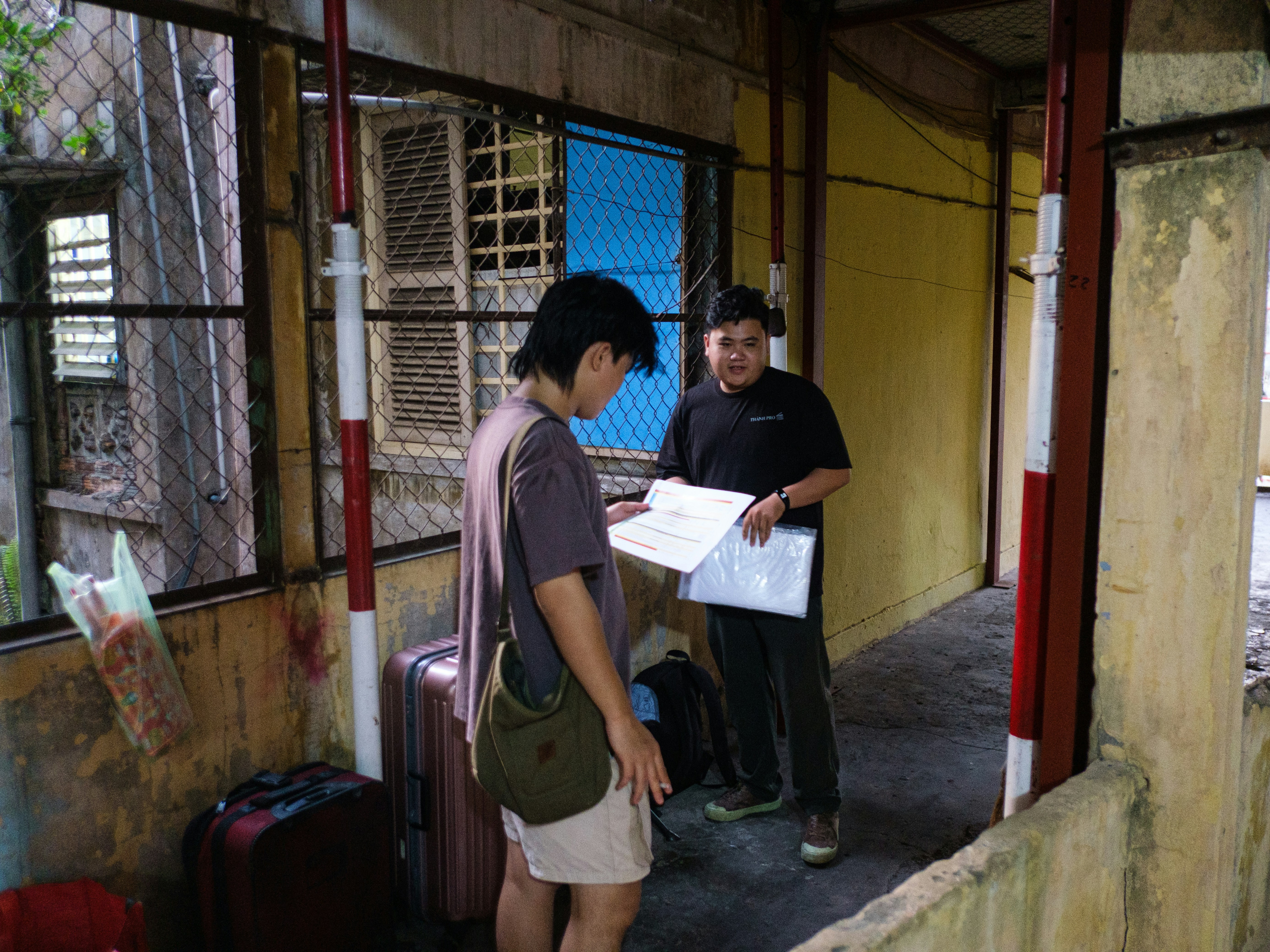 Two people discussing documents in a worn hallway with luggage
