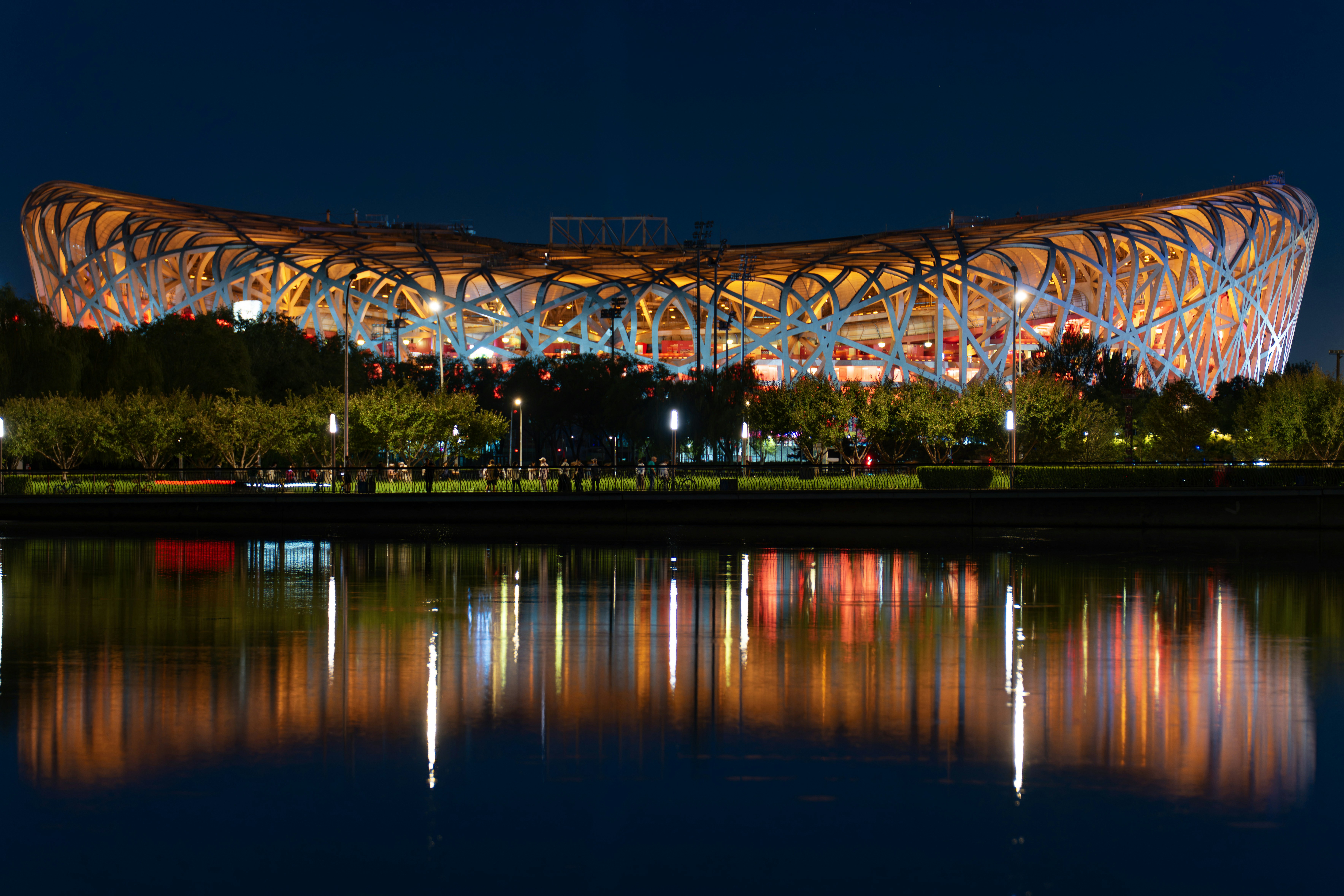 Illuminated bird's nest stadium reflected in water at night.