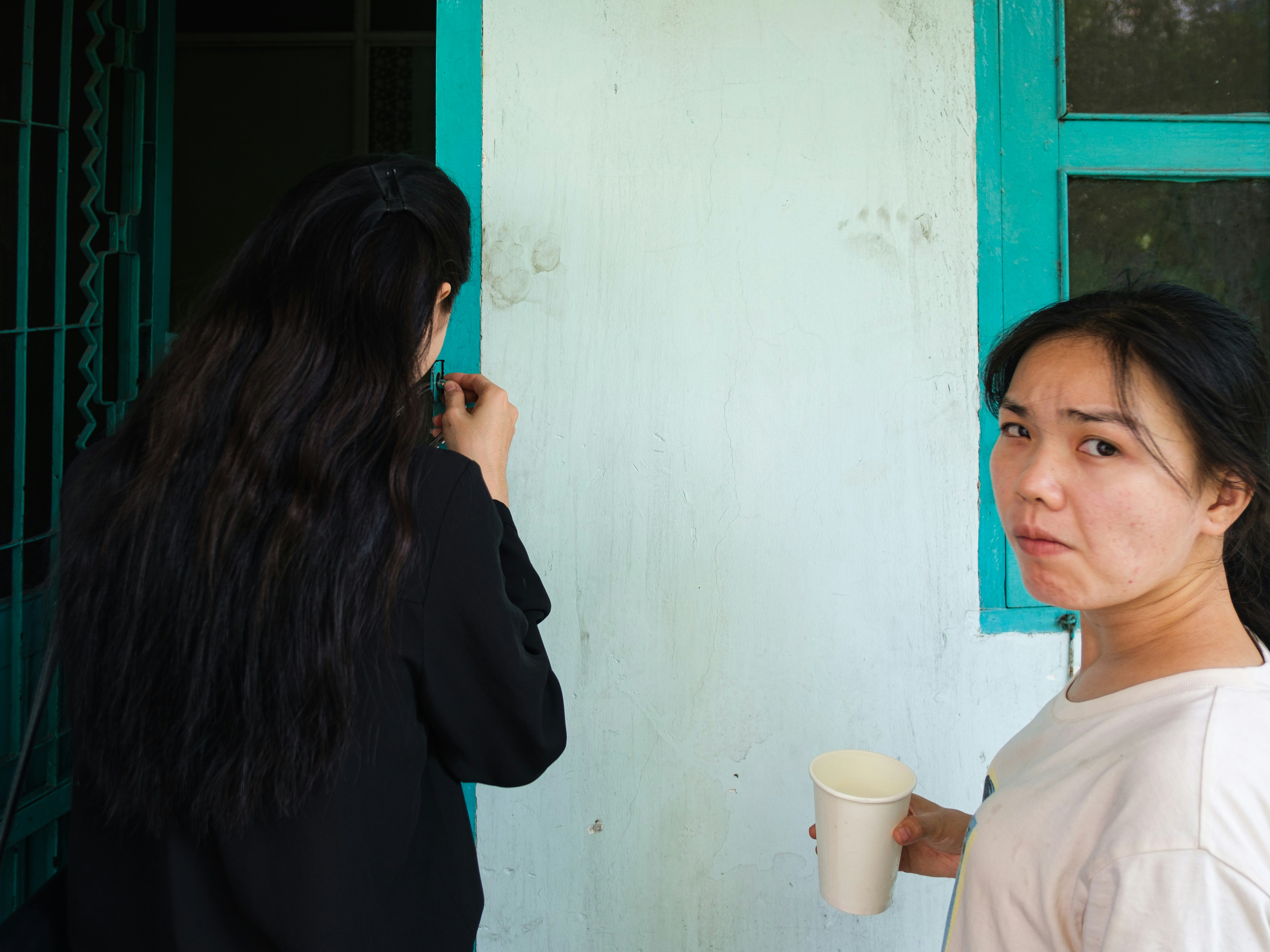 Woman with cup looks at camera, another woman faces wall