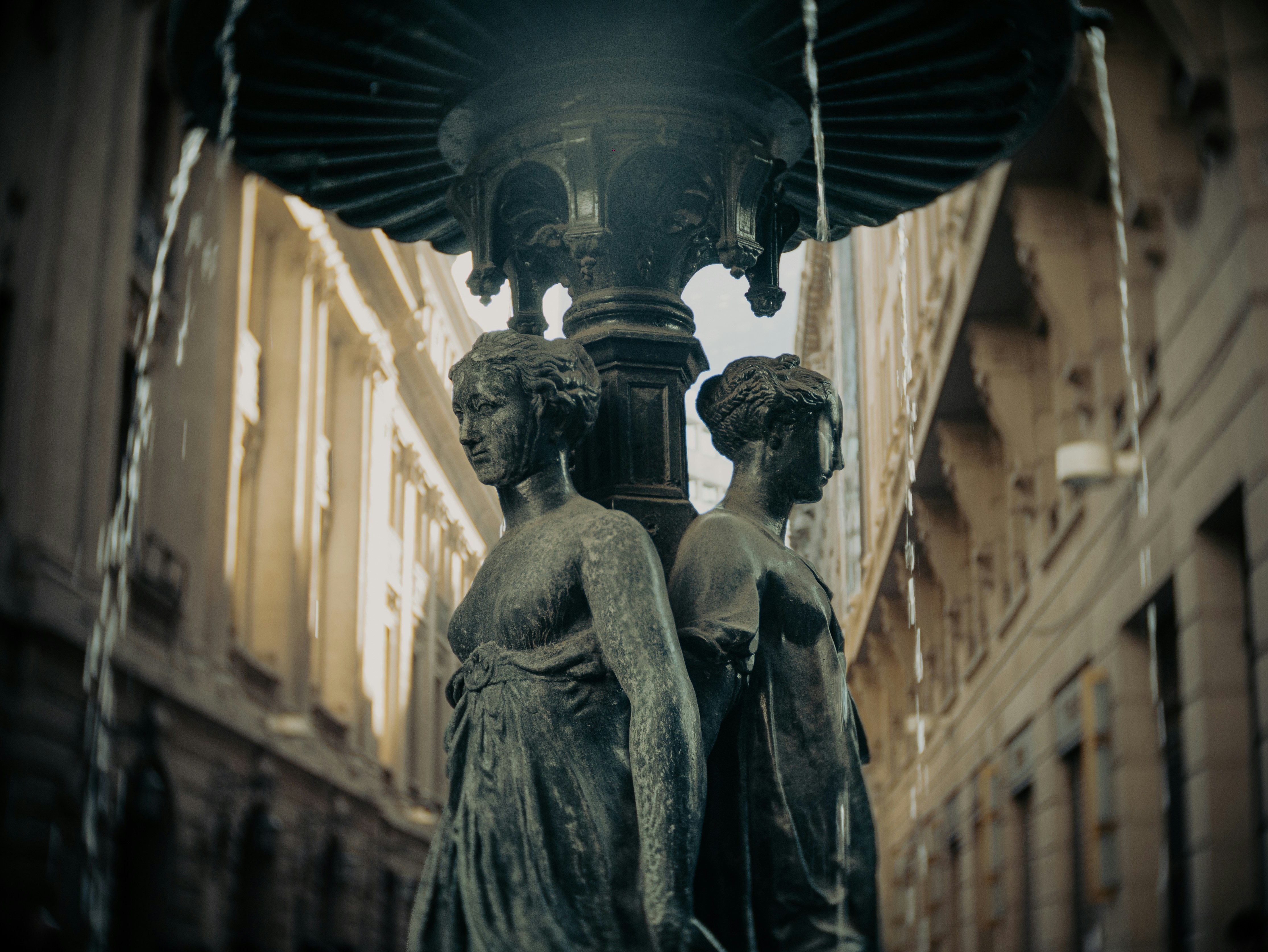 Two classical female statues adorn a dark, ornate city fountain.