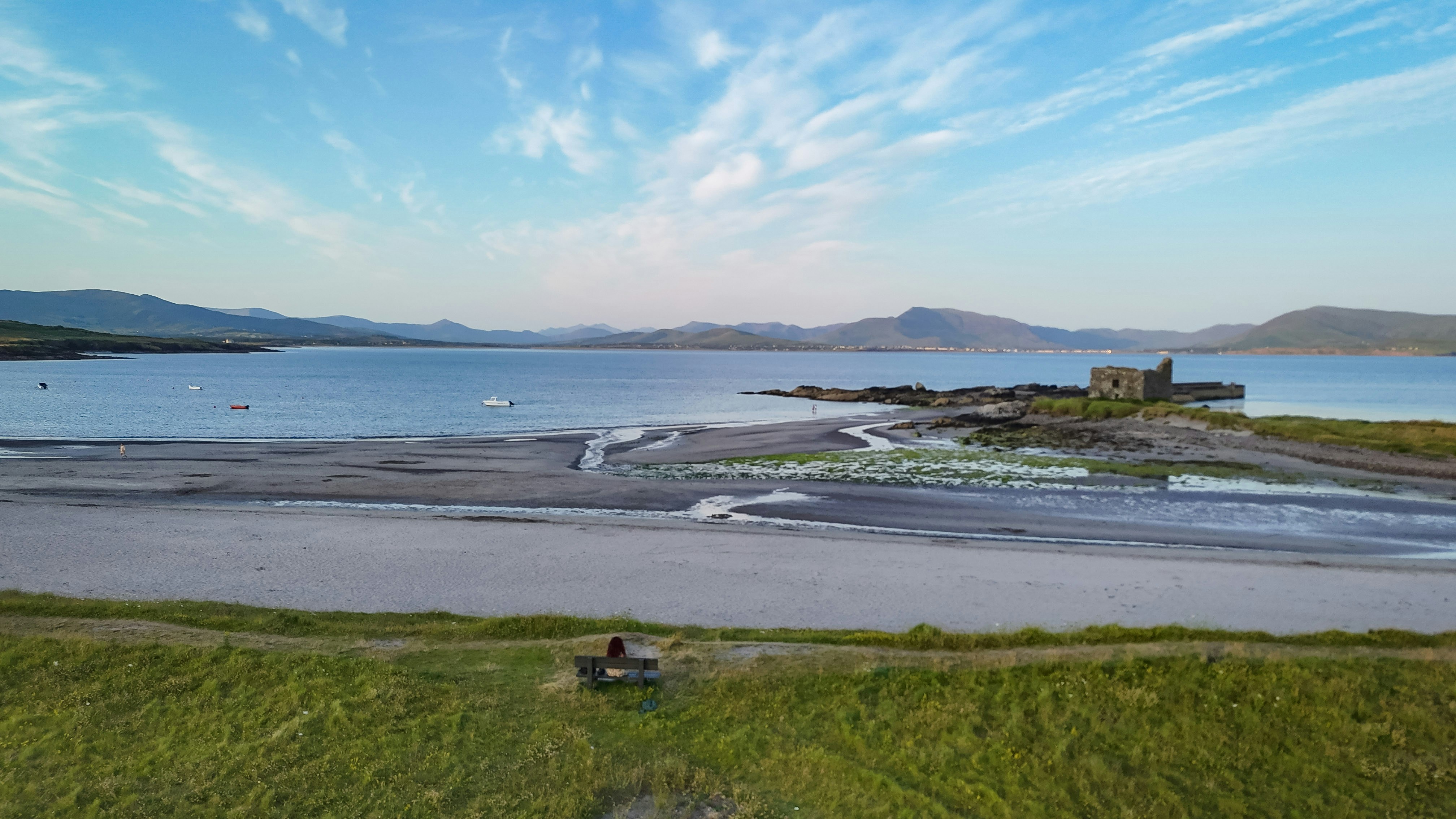 Person on bench overlooking coastal landscape with ruins and mountains.