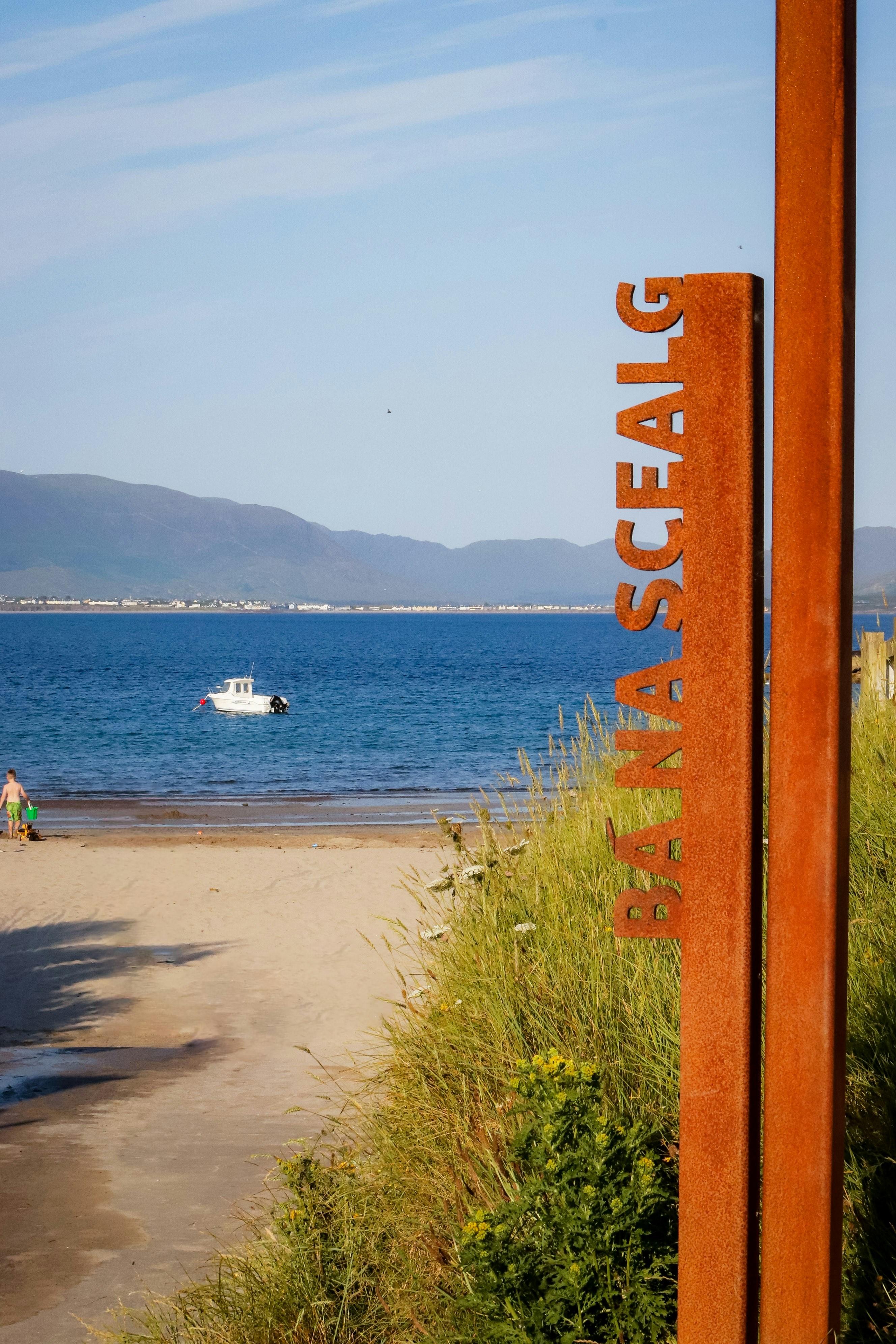 Rusty sign beside a beach, ocean, and distant mountains.