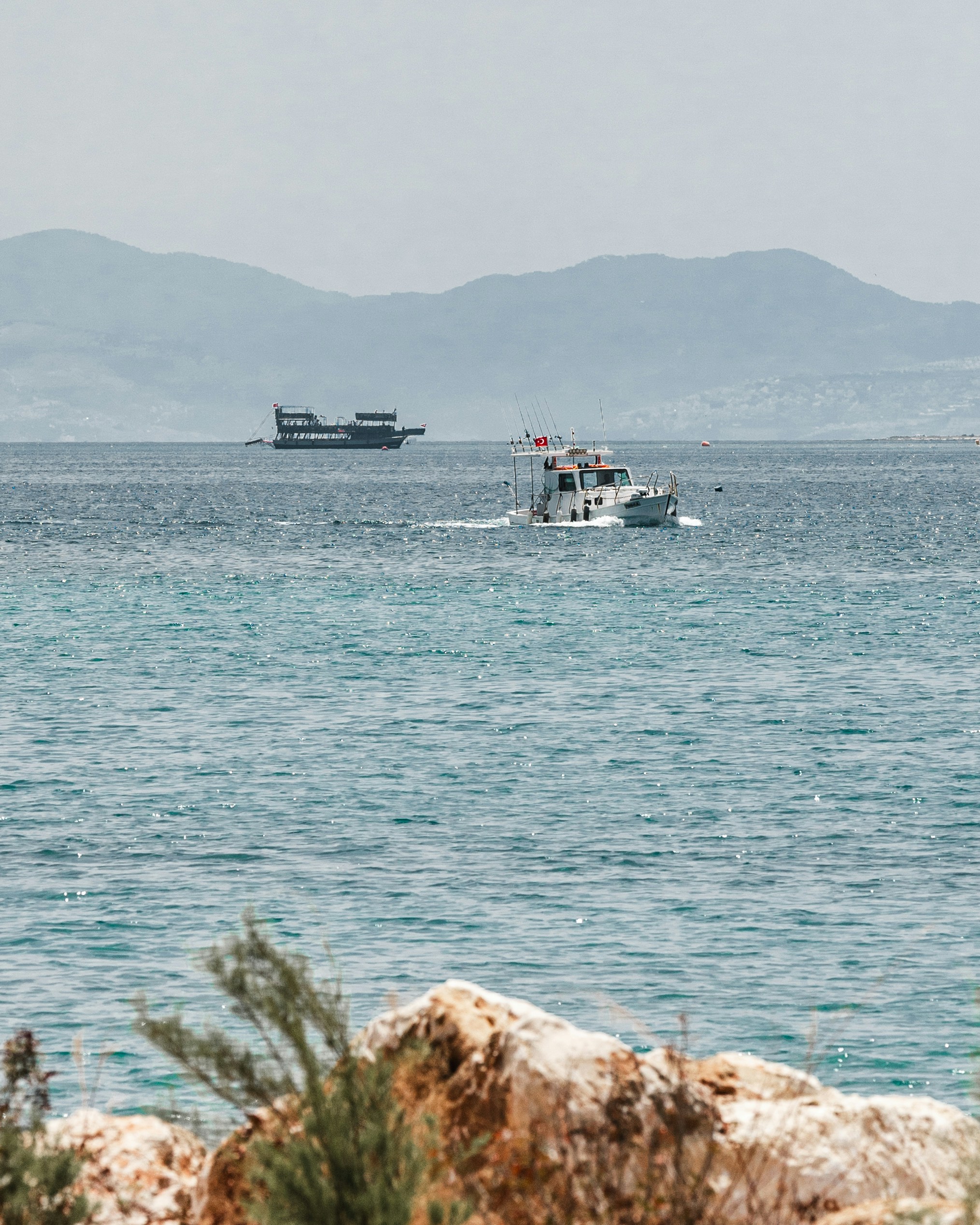 Boats on the sea with mountains and rocky foreground.