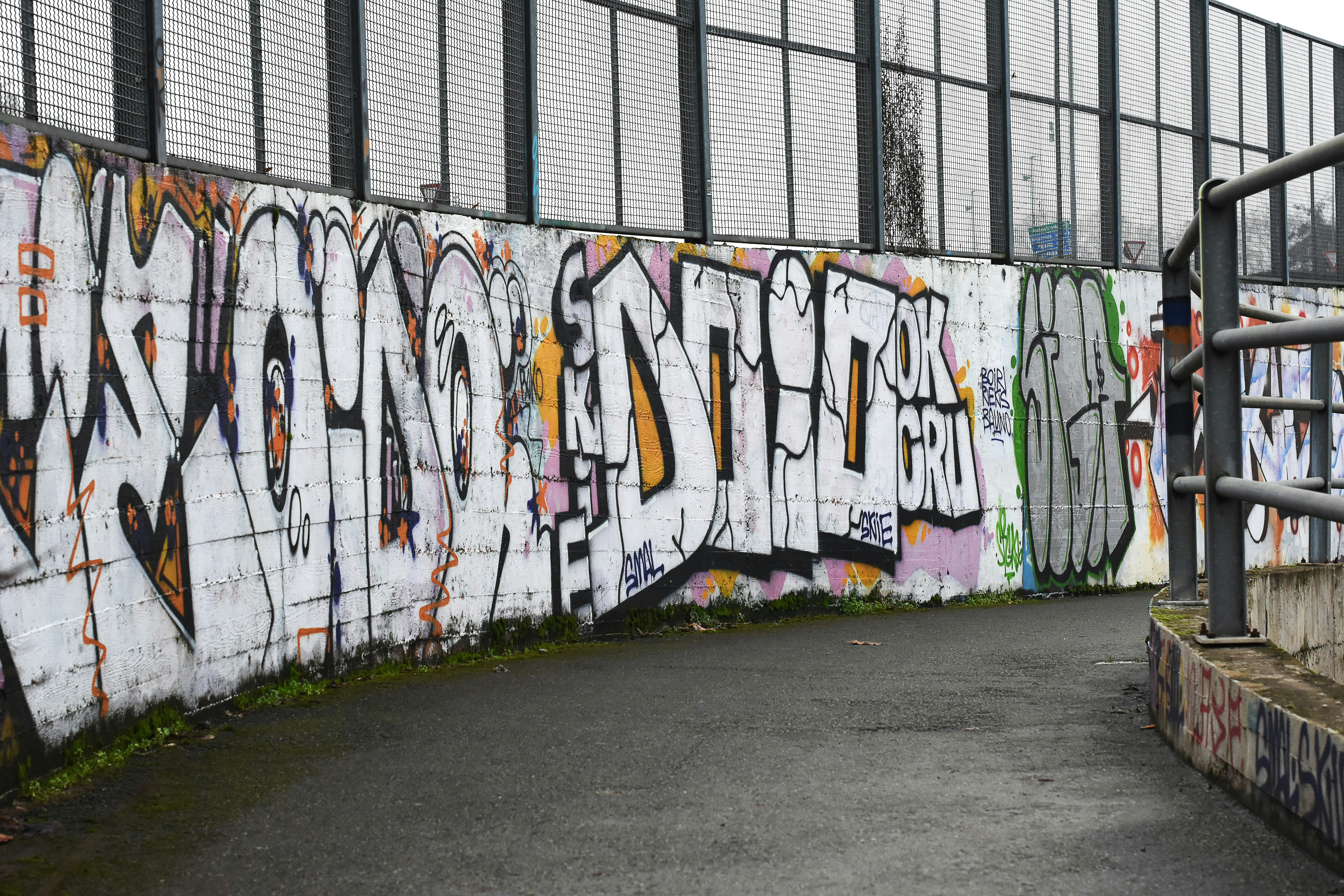 Graffiti-covered wall alongside a paved path under a metal fence.
