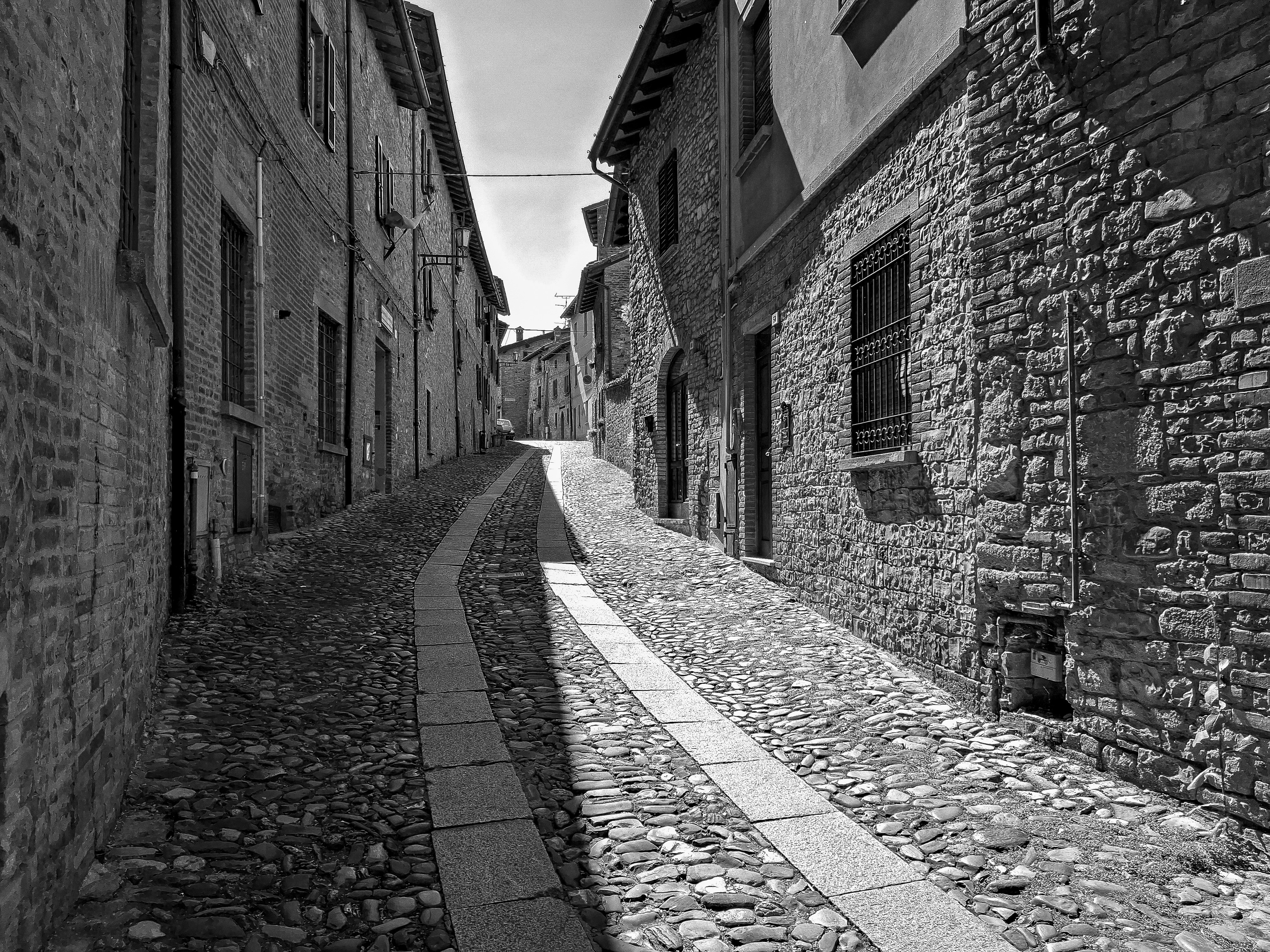 Strada monocromatica e sinuosa in ciottoli tra antichi edifici in pietra.