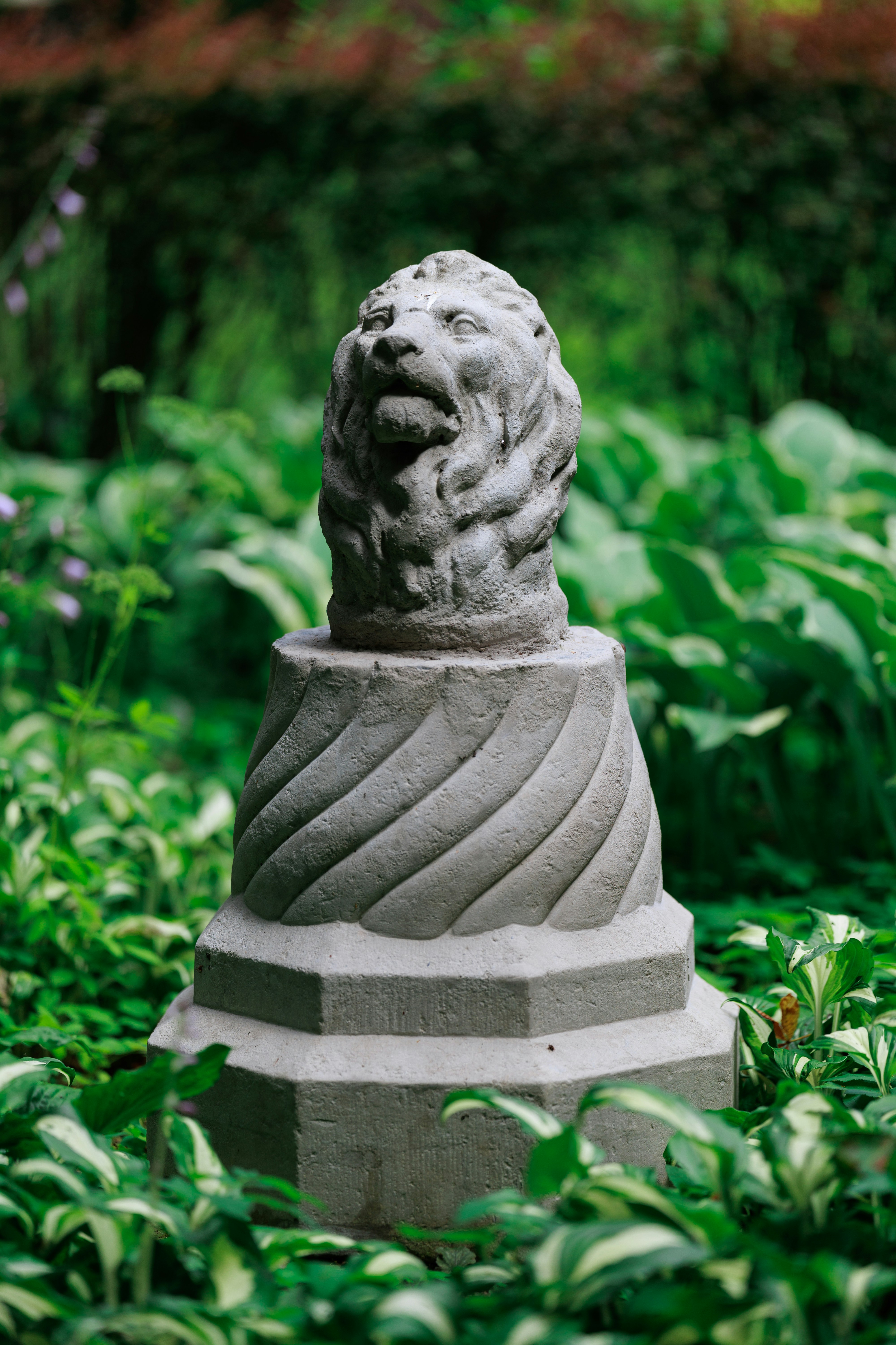 Estatua de cabeza de león de piedra sobre un pedestal entre follaje verde.