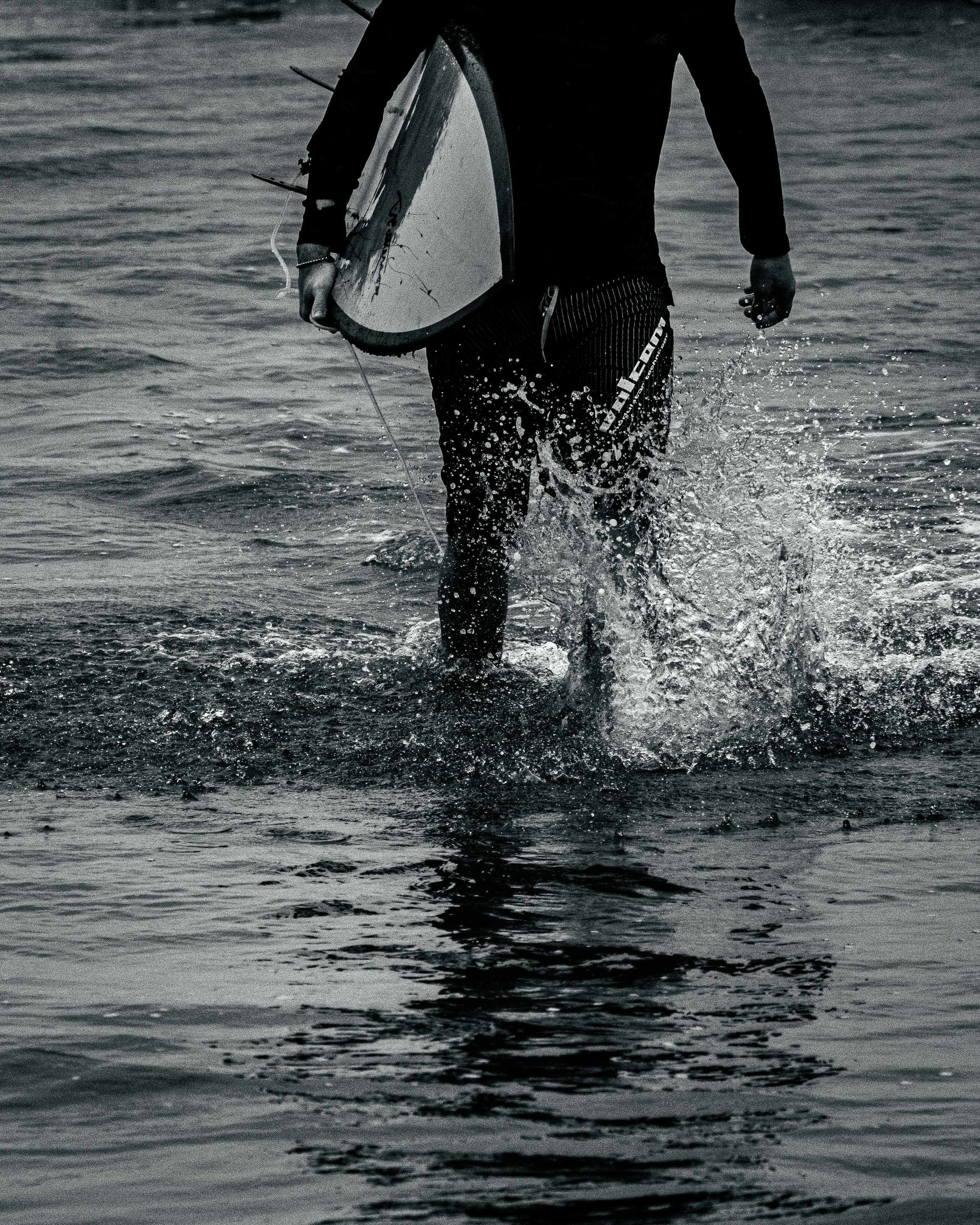 Surfer in wetsuit walks through ocean water carrying a surfboard.