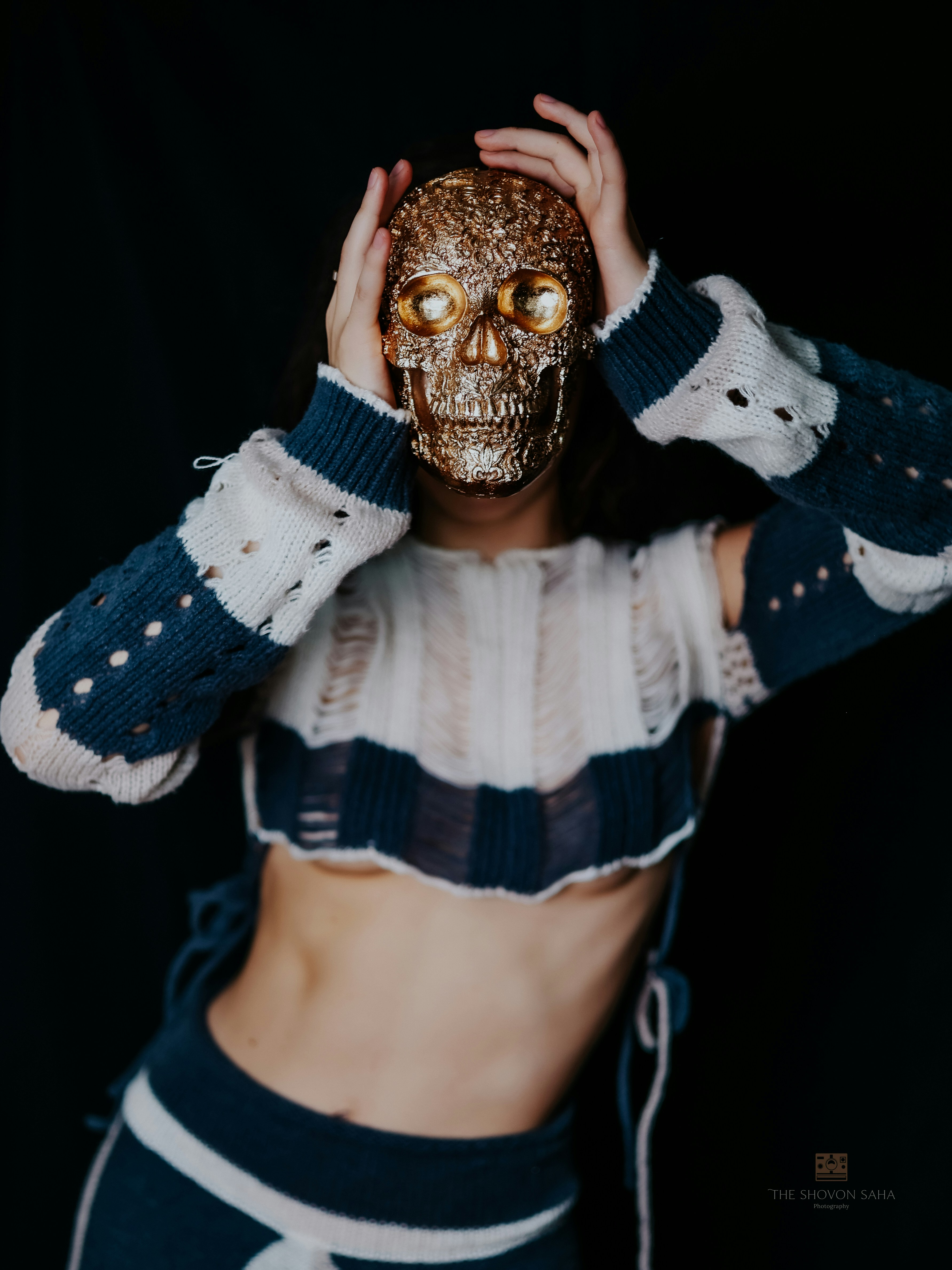 A person holding a gold skull mask over their face wearing a distressed sweater