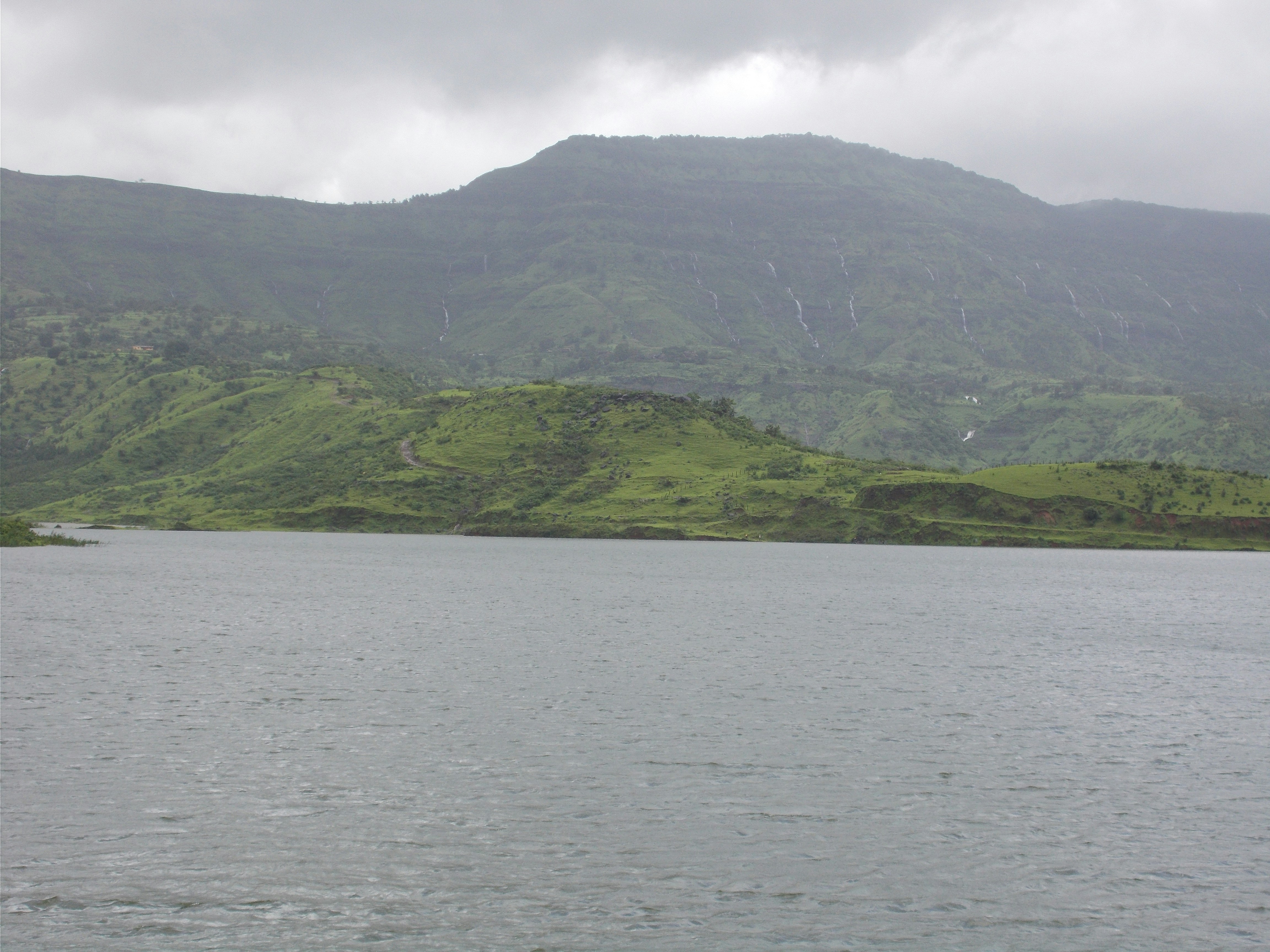A tranquil lakeside scene with calm water and distant green hills under cloudy skies. The image emphasizes the broad landscape with soft, diffused light and a gentle horizon.