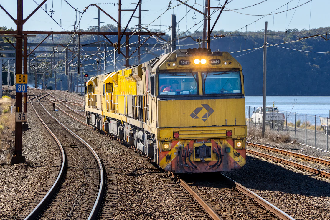 A yellow freight locomotive runs beneath overhead power lines