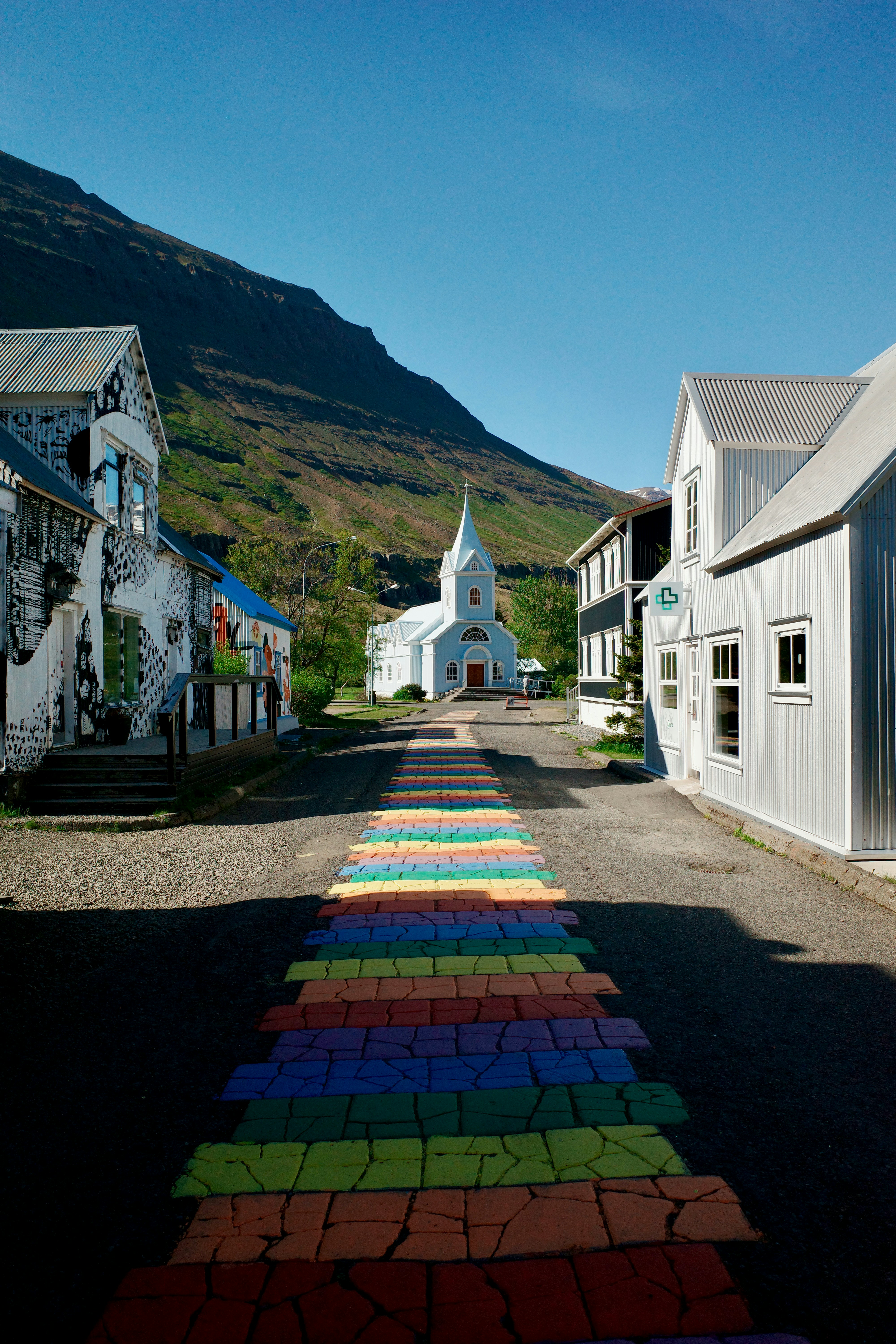 Une rue peinte d’arc-en-ciel approche d’une église blanche avec des montagnes derrière.