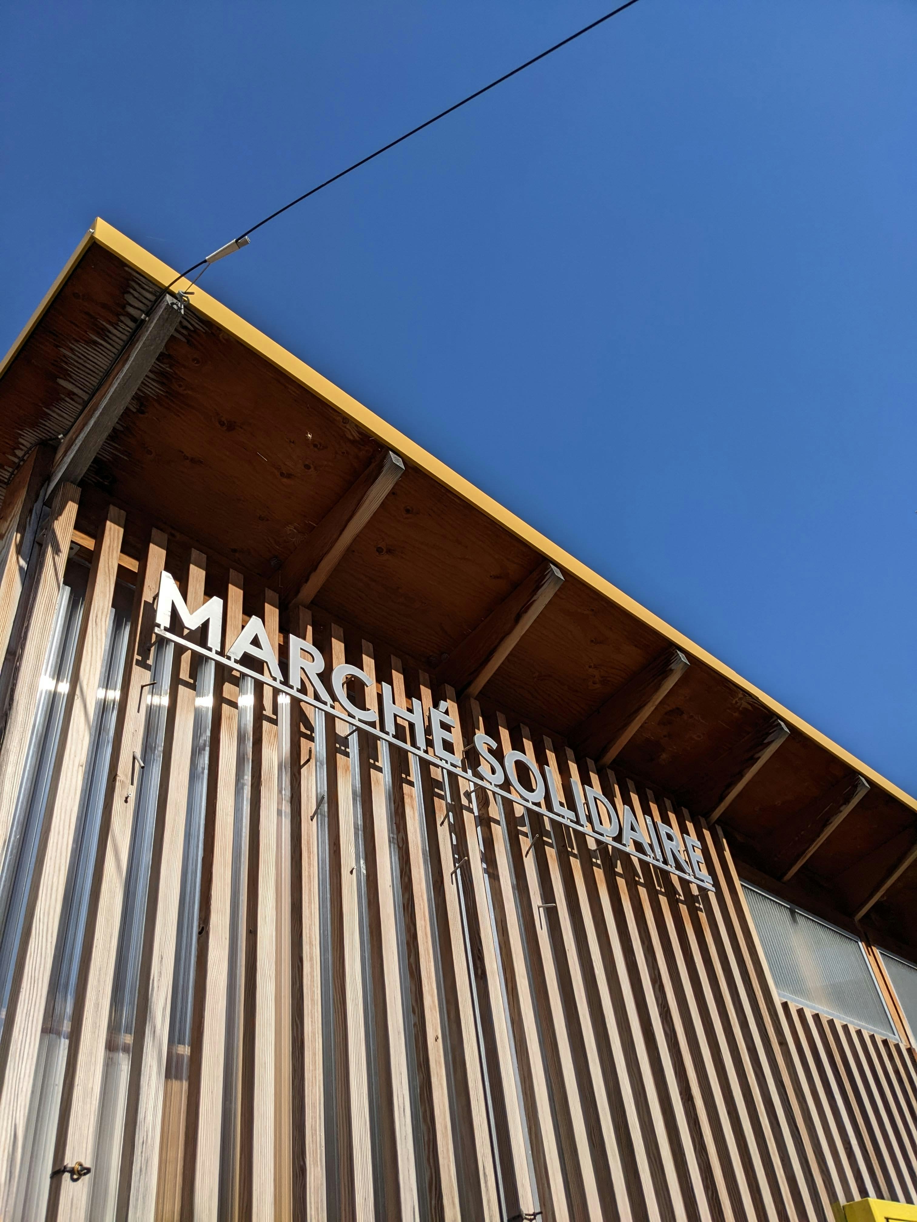Wooden building with 'marche solidaire' sign against blue sky.