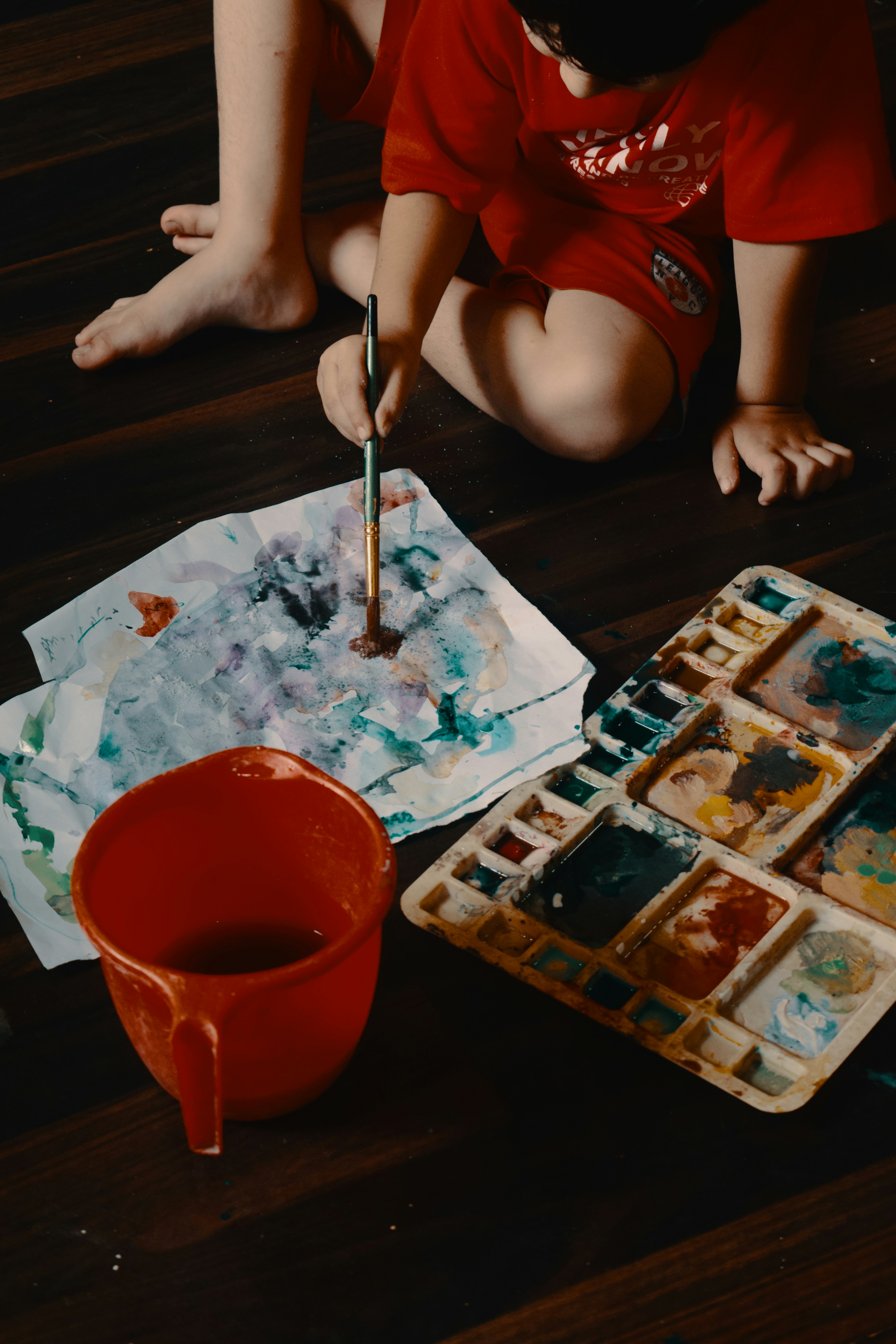 A child painting with watercolors on a wooden floor.