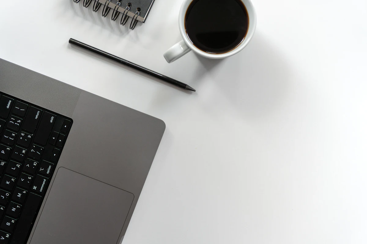 Laptop, buku catatan, pena, dan secangkir kopi tertata di meja kerja pemilik toko