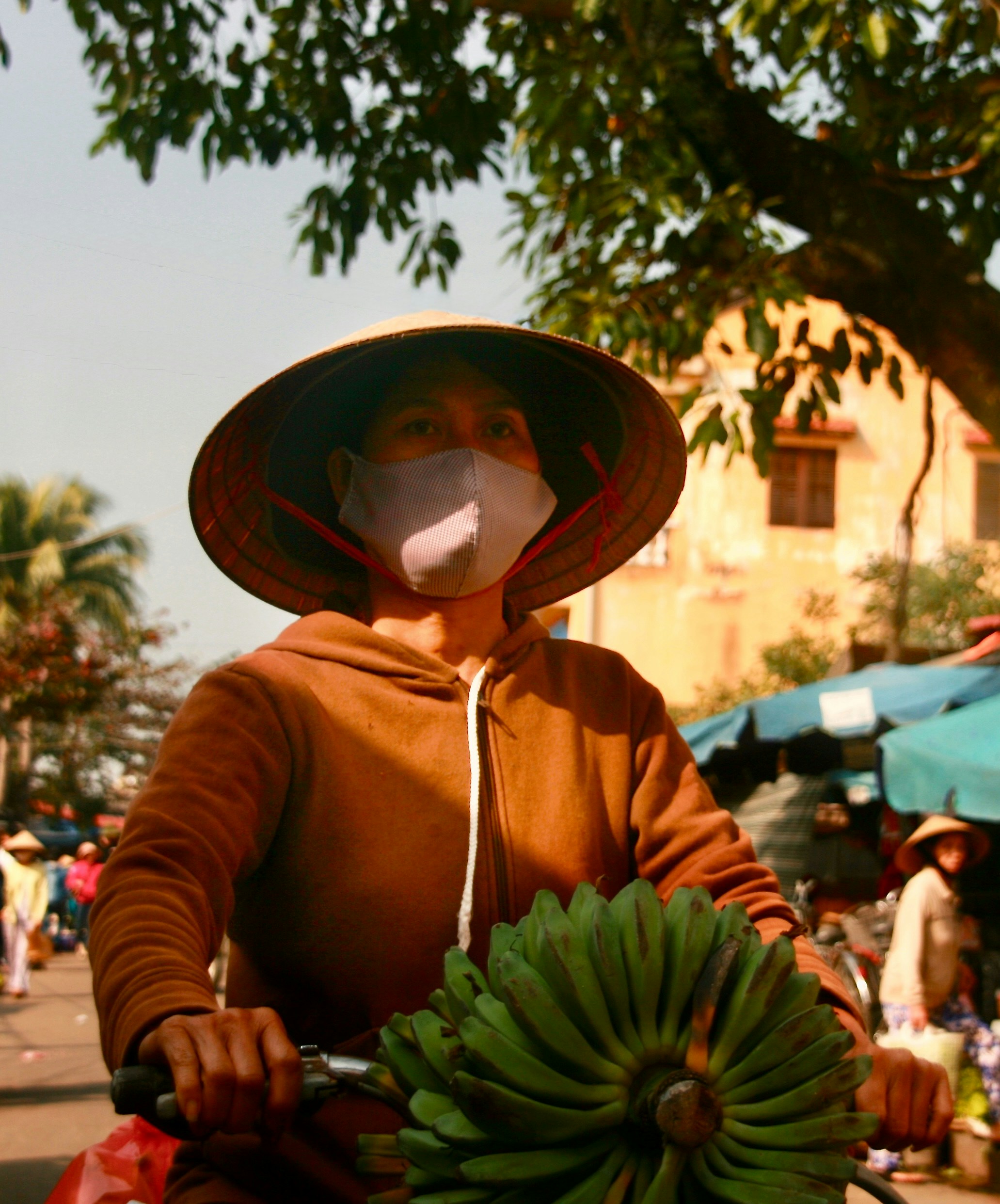 Woman wearing conical hat and mask cycling with bananas.